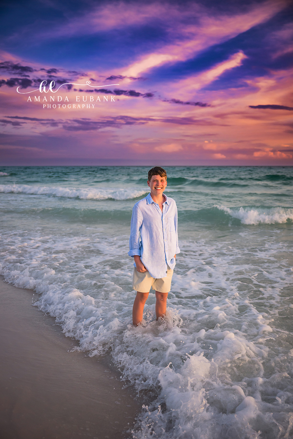 Seaside FL Tween Boy in Gulf Surf | Twilight Beach Portrait Tween boy in a light blue linen button-up and cream shorts smiles while standing in the foamy Seaside, FL Gulf surf beneath a vivid pink and purple twilight sky.