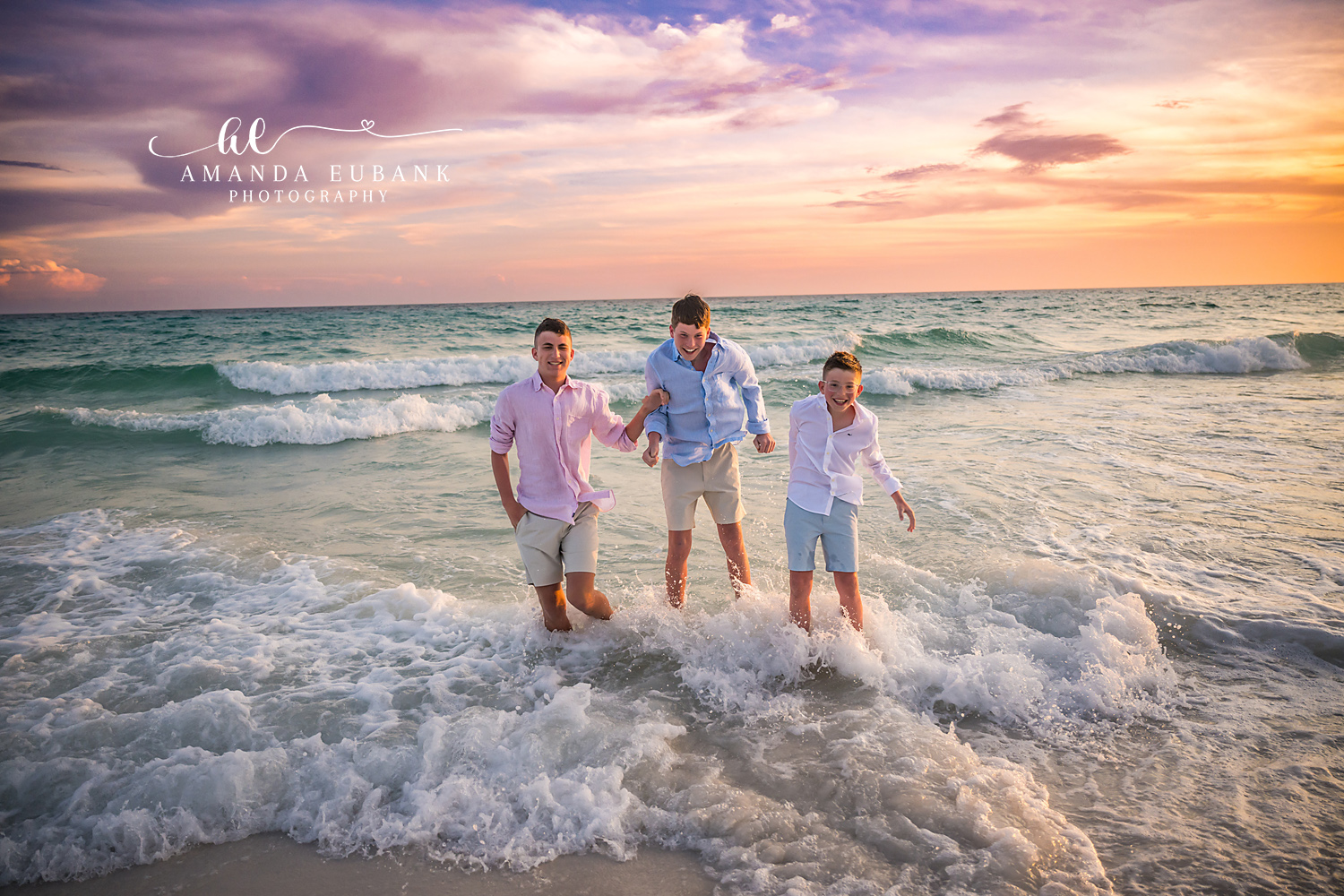 Three brothers in pink, light blue, and white linen splash through the foamy Seaside, FL Gulf surf together as a glowing peach and orange sunset lights up the sky.