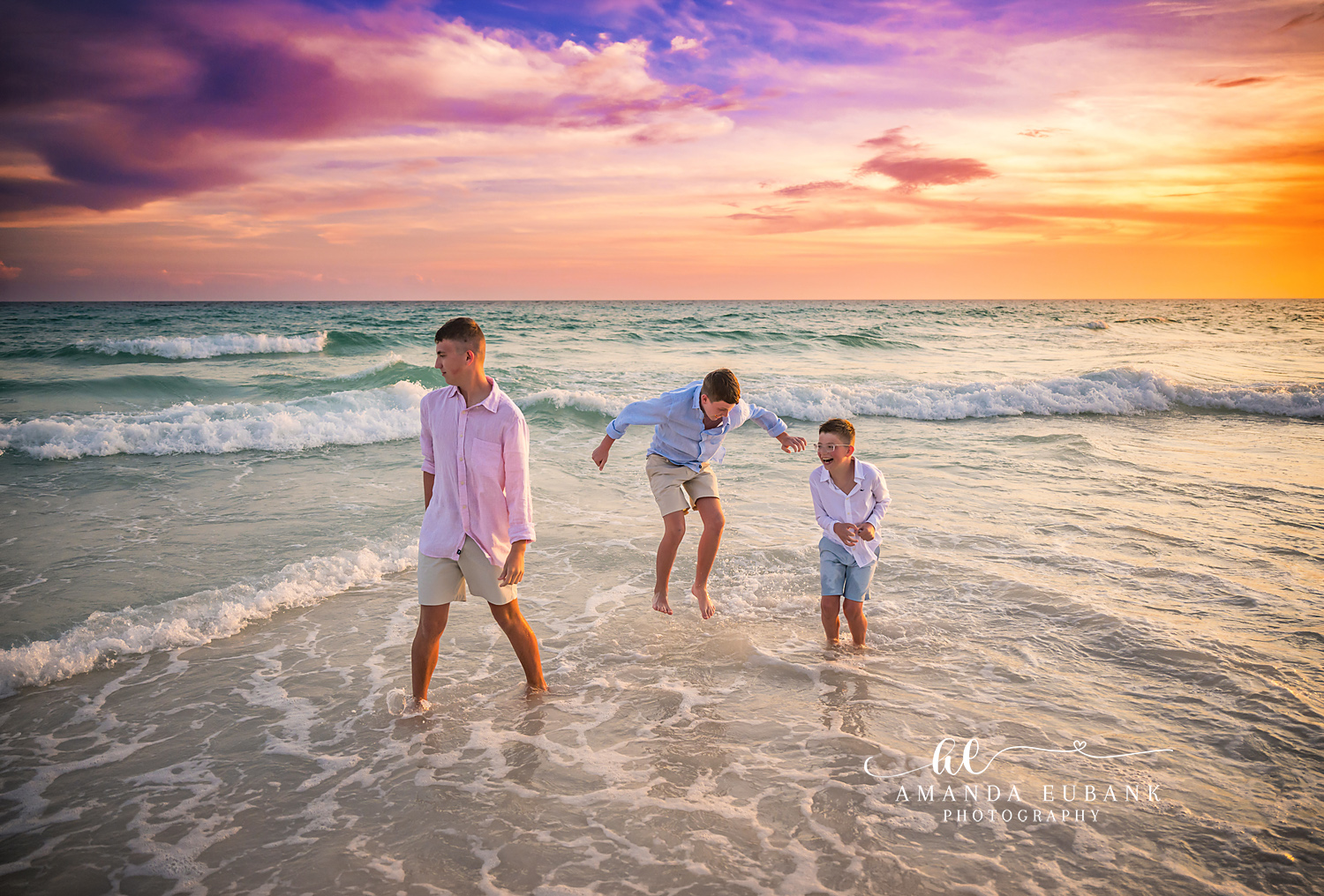Seaside FL Brothers Playing in Surf | Candid Sunset Beach Moment Three brothers laughing and jumping through the foamy Seaside, FL Gulf surf at sunset, coordinated in pink, light blue, and white linen.