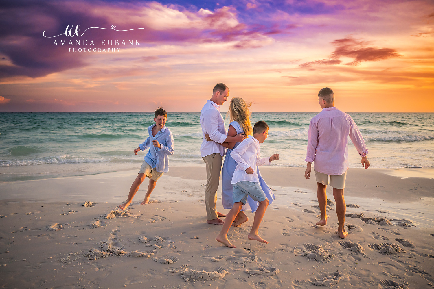Seaside FL Candid Family Sunset Moment | Parents Embrace as Boys Play Mom and dad embrace as their three sons run and play around them along the Seaside, FL shoreline at golden hour, with a brilliant orange sunset over the Gulf.