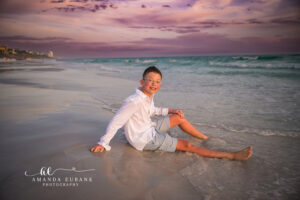 Smiling young boy with glasses sits on the wet sand at the Seaside, FL surf line in a white linen button-up and light blue shorts beneath a soft purple twilight sky.