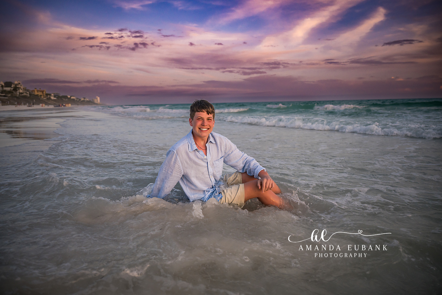 Seaside FL Boy Sitting in Gulf Surf | Twilight Beach Portrait Smiling tween boy sits in the rolling Seaside, FL Gulf surf in a light blue button-up and khaki shorts as soft lavender twilight clouds drift over the Emerald Coast horizon.