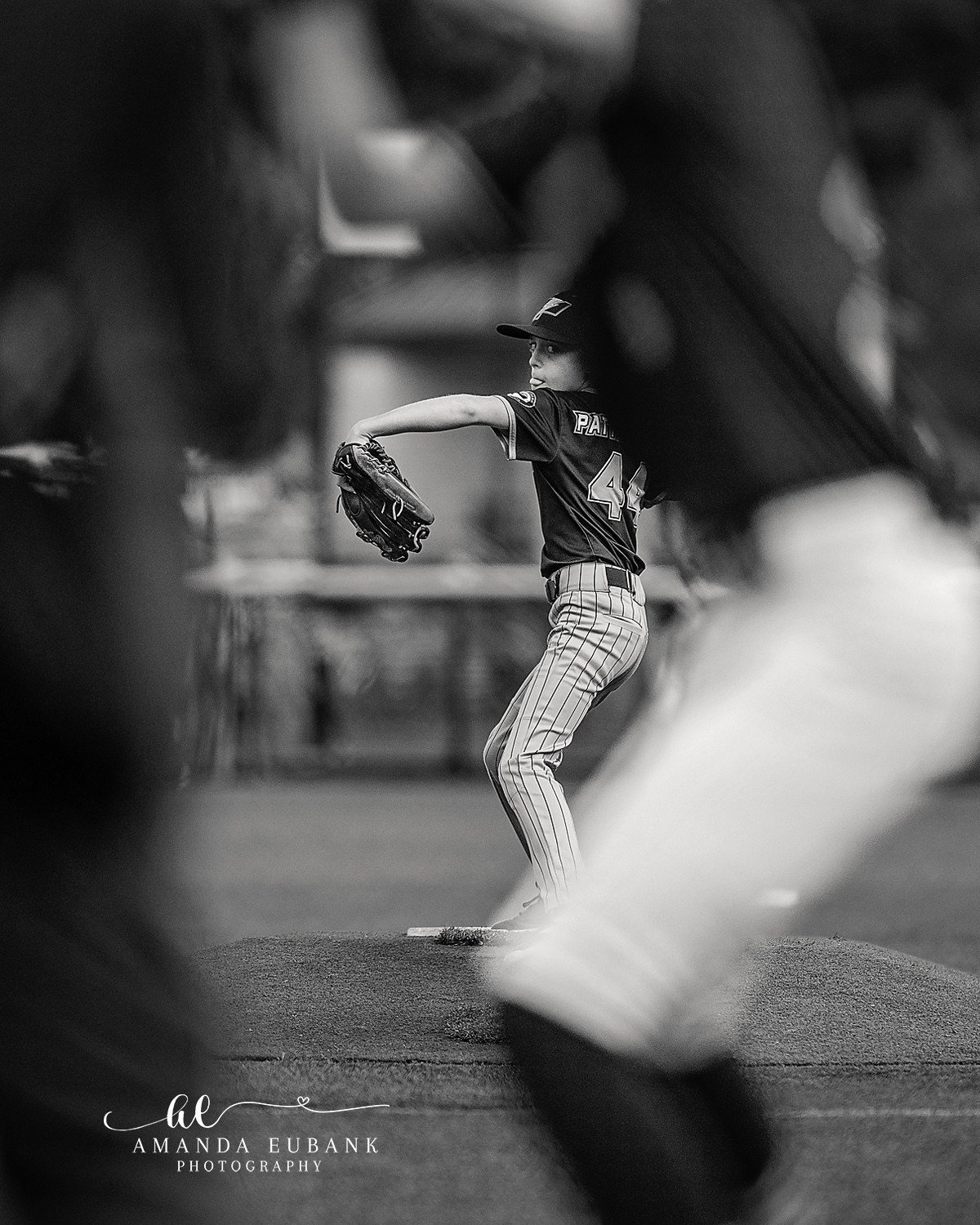Pitcher Through the Bat Frame | Hoover AL Travel Baseball Photographer Black and white creative frame showing a Power Kilcrease 11U pitcher mid-windup on the mound, viewed through the motion-blurred swinging bat of a foreground teammate at a Hoover, Alabama tournament.
