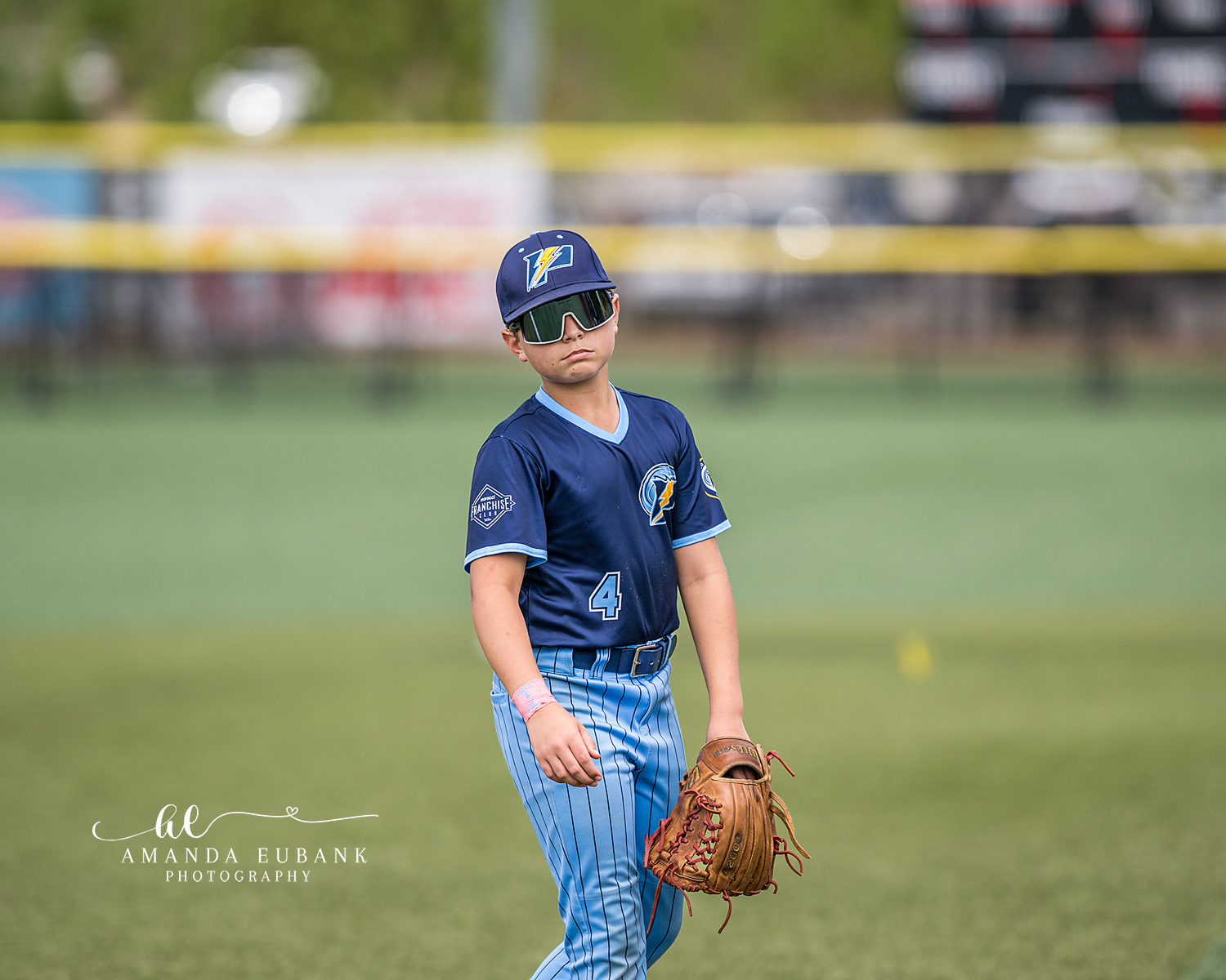 Player Number 4 Sunglasses Portrait | Hoover AL Travel Baseball Photographer Power Kilcrease 11U player number 4 walks the field in sport sunglasses, navy jersey, and light blue pinstripe pants holding his tan glove during a Hoover, Alabama travel baseball tournament.