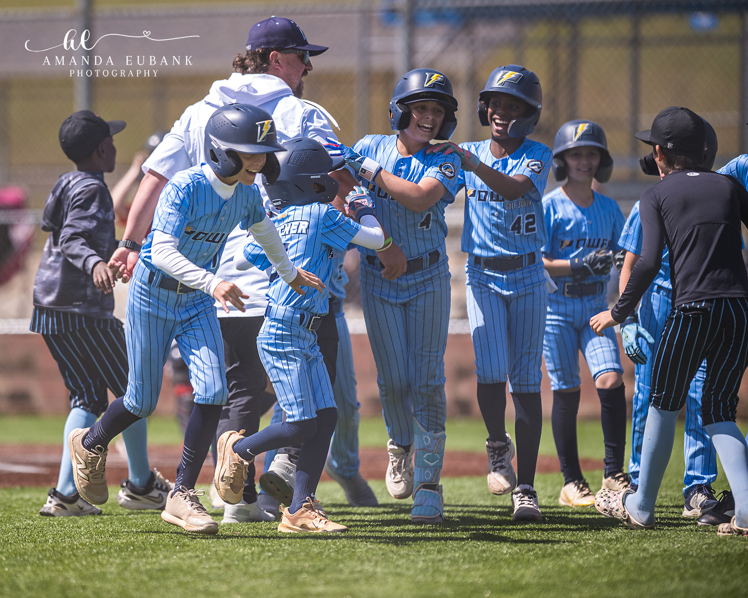 Smiling Power Kilcrease 11U players in light blue Power jerseys and pinstripe pants celebrate together on the infield, jersey number 42 at the center, during a Hoover, Alabama tournament game.