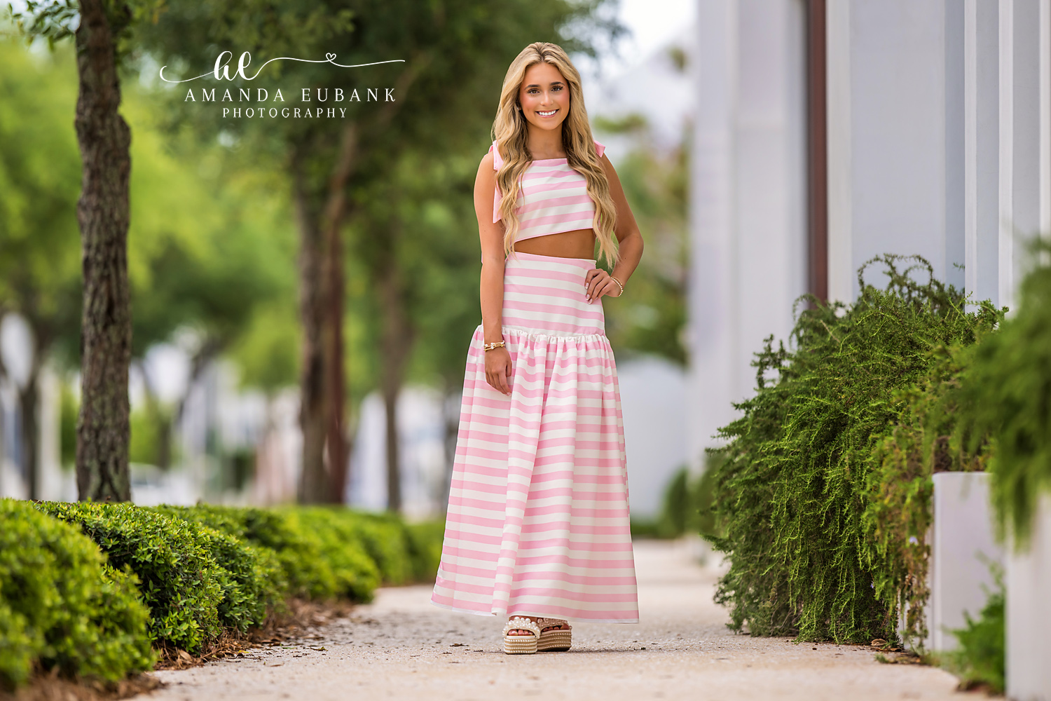 Senior Portrait in Pink Striped Set on a Garden Path | Photographer in Alys Beach Senior in a pink and white striped two-piece set with tiered maxi skirt on a sandy garden path lined with hedges and palms, by Photographer in Alys Beach Amanda Eubank.