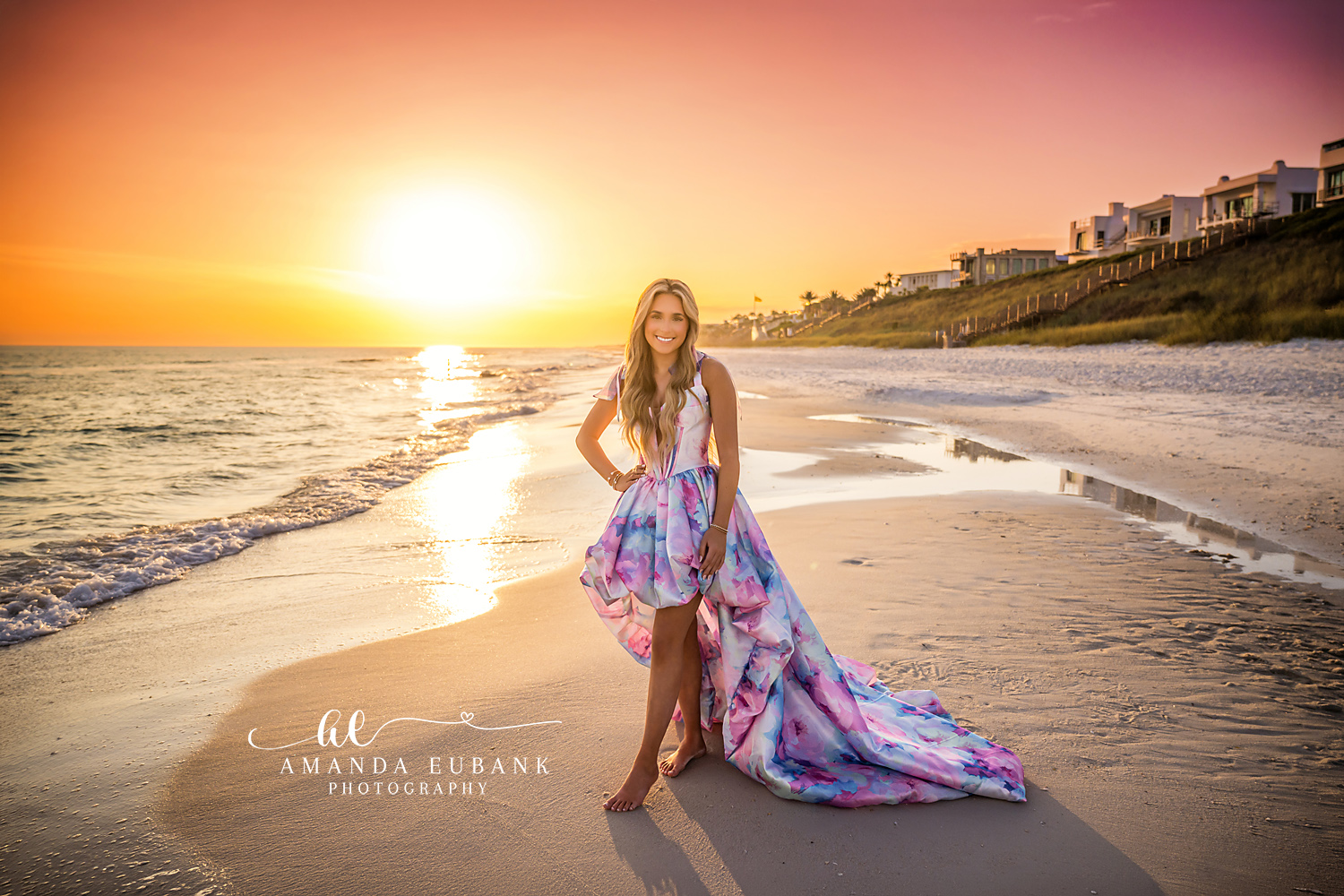 Sunset Senior Portrait on the Shoreline with Dune Homes | Photographer in Alys Beach Senior in a pastel watercolor high-low gown standing barefoot on the wet sand at sunset with the dune homes behind her, by Photographer in Alys Beach Amanda Eubank.