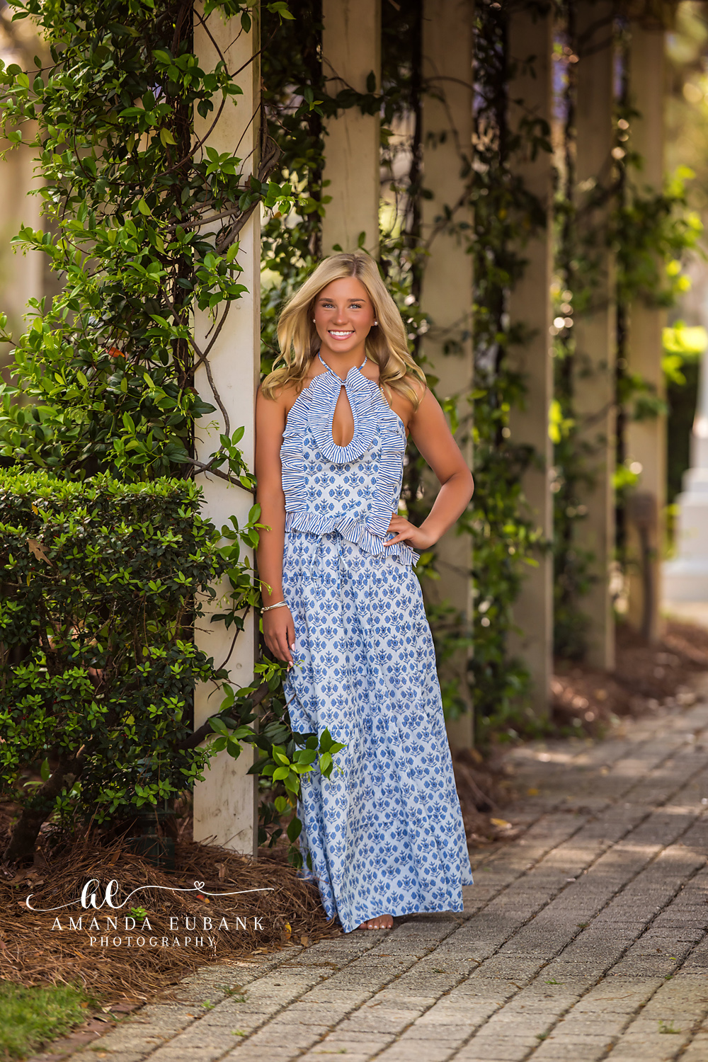Rosemary Beach Brick Walkway Senior Portrait | 30A Senior Photographer Senior girl in blue and white halter maxi dress on a brick walkway beside a vine-wrapped column in Rosemary Beach FL