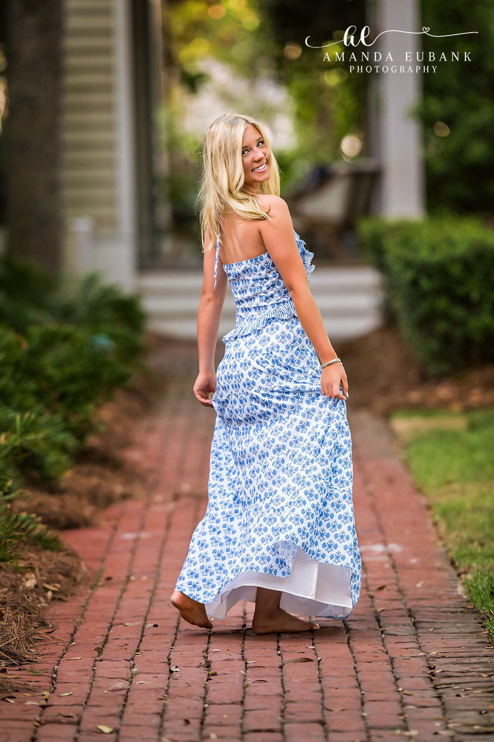 Rosemary Beach Look-Back Senior Portrait | 30A Senior Photographer Senior girl glancing back over her shoulder while walking down a Rosemary Beach brick paver street in a blue and white halter maxi dress