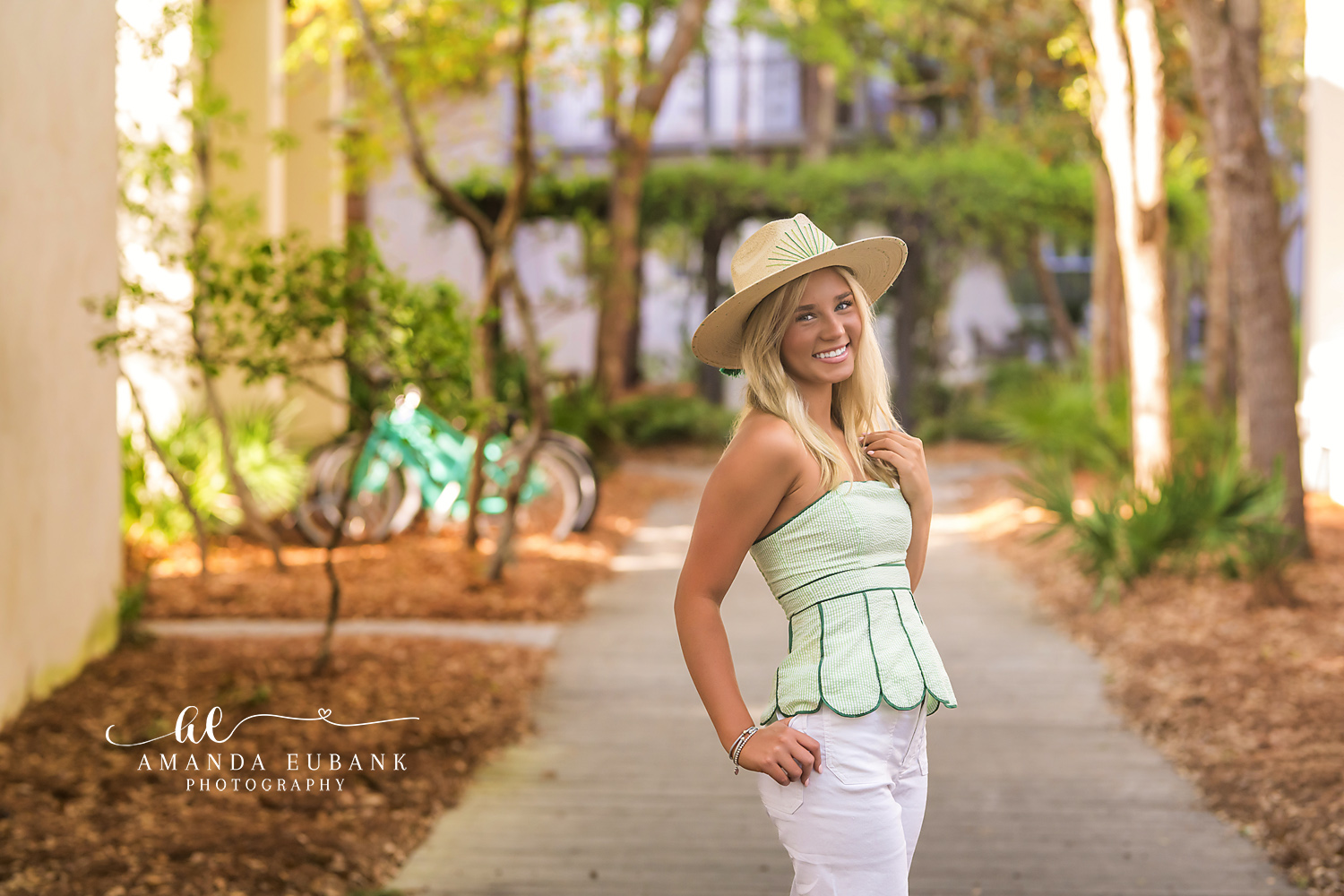 Rosemary Beach Beach Cruiser Senior Portrait | 30A Senior Photographer Senior girl in sage green top and straw hat smiling on a Rosemary Beach lane with pastel cottages and teal beach cruisers behind her