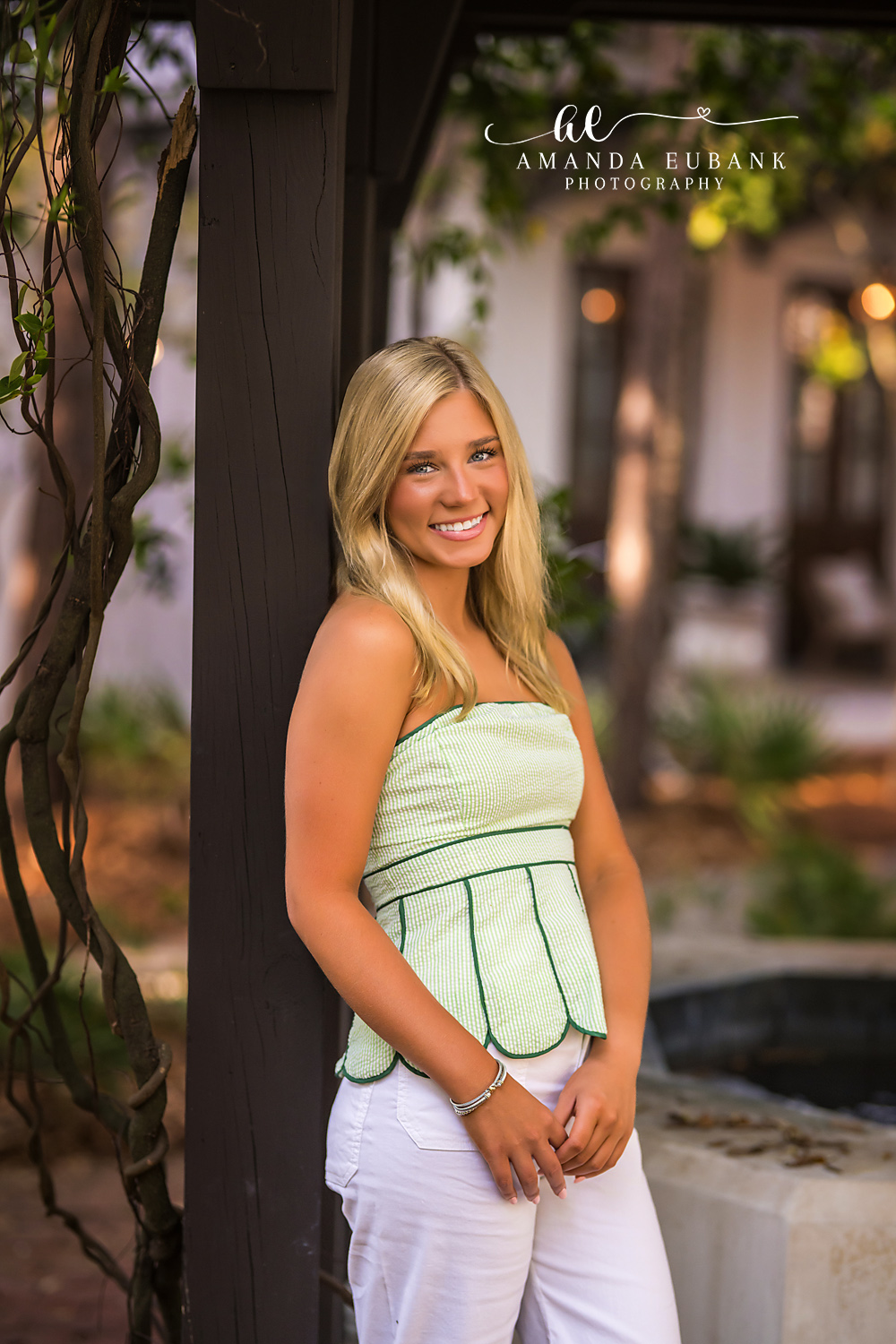 Rosemary Beach Pergola Post Senior Portrait | 30A Senior Photographer Senior portrait of a smiling girl in a sage green scalloped top beside a vine-wrapped pergola post in a Rosemary Beach courtyard
