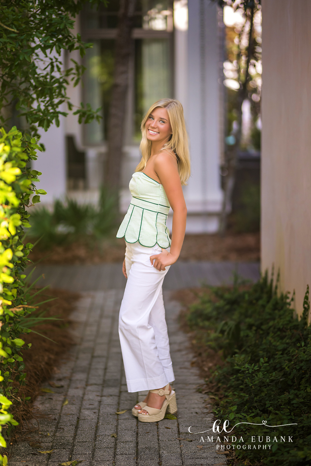 Rosemary Beach Garden Lane Senior Portrait | 30A Senior Photographer Senior girl in sage green top and white pants smiling on a brick paver garden lane between Rosemary Beach cottages