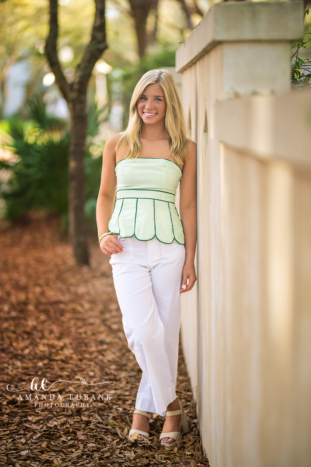 Rosemary Beach Stucco Wall Senior Portrait | 30A Senior Photographer Senior girl in a sage green top and white pants standing beside a cream stucco wall under live oaks in Rosemary Beach FL