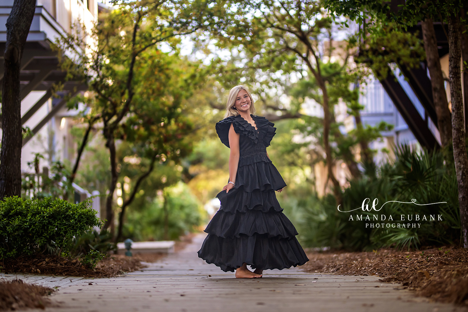 Rosemary Beach Boardwalk Black Gown Senior Portrait | 30A Senior Photographer Senior girl in a black tiered ruffle gown holding her skirt on a Rosemary Beach boardwalk framed by live oak trees