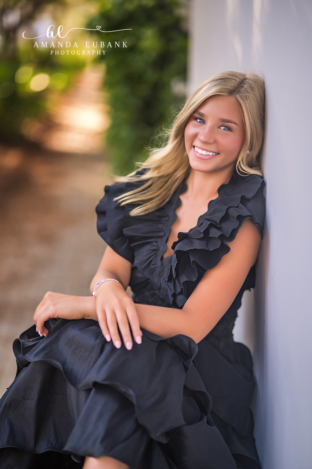 Rosemary Beach Seated Senior Portrait | 30A Senior Photographer Seated senior portrait of a smiling girl in a black ruffle gown leaning on her knee against a soft golden bokeh background in Rosemary Beach FL