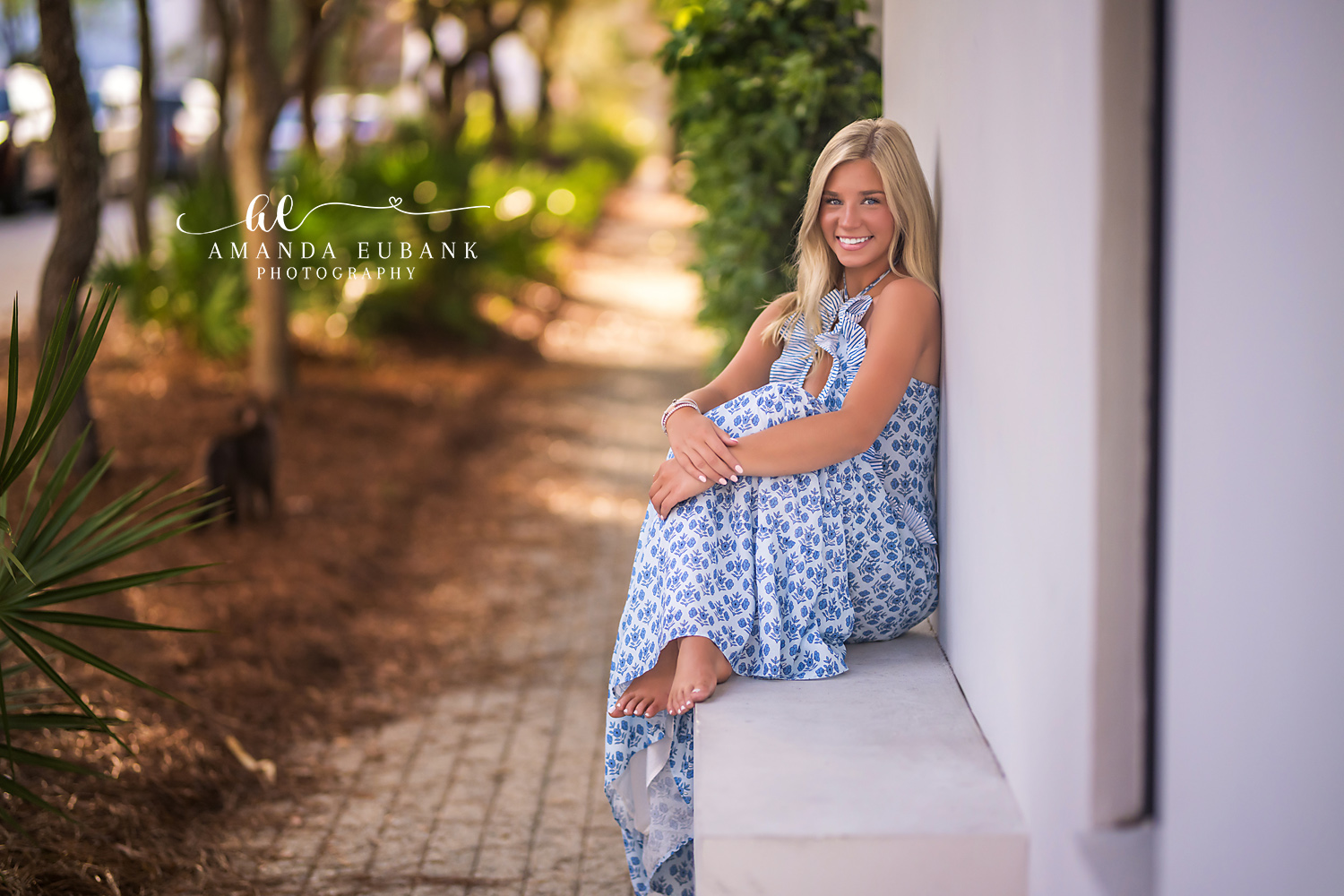 Rosemary Beach Garden Wall Senior Portrait | 30A Senior Photographer Senior girl in blue and white halter maxi dress sitting barefoot on a low white wall along a Rosemary Beach garden path
