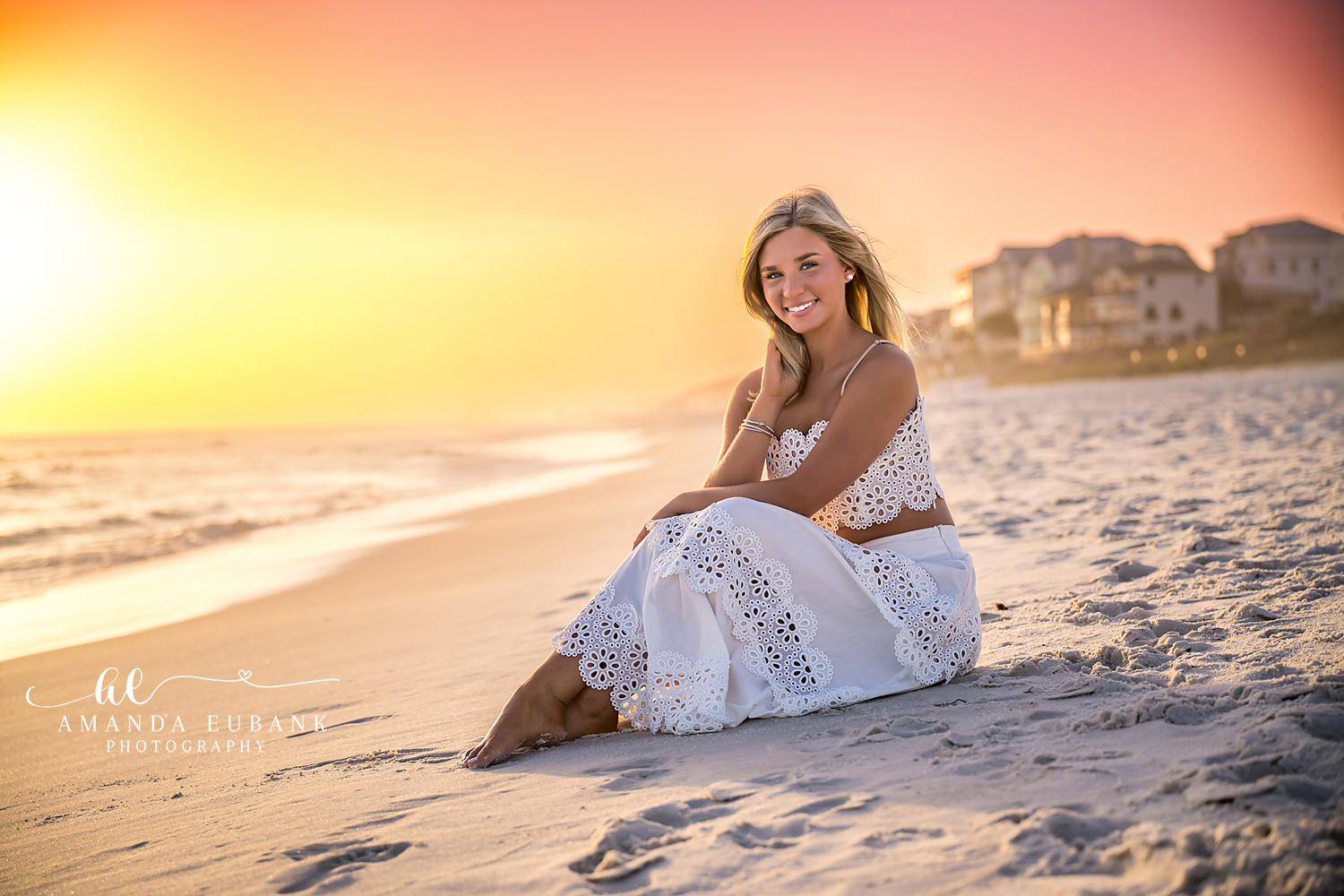 Rosemary Beach Beachfront Sunset Senior Portrait | 30A Senior Photographer Senior girl in white eyelet two-piece sitting barefoot on the Rosemary Beach sand at sunset with pastel 30A cottages on the horizon