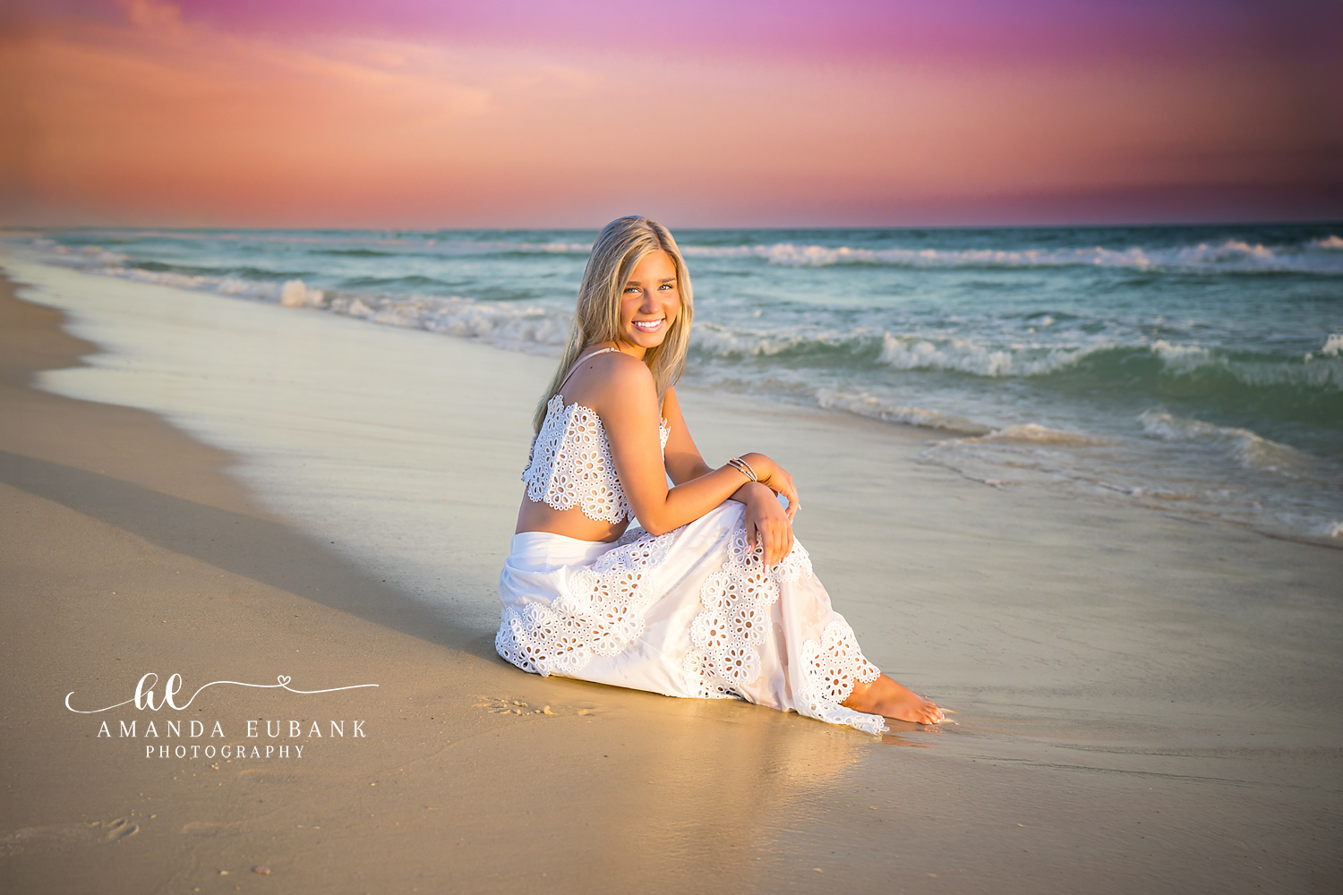 Rosemary Beach Pastel Sunset Shoreline Senior Portrait | 30A Senior Photographer Senior girl in white eyelet two-piece sitting barefoot on the Rosemary Beach shoreline at sunset with pastel pink and teal sky behind her