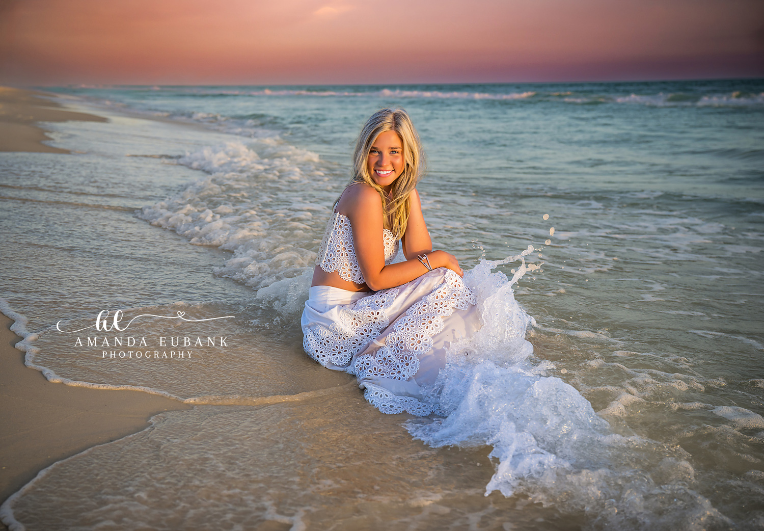 Senior girl kneeling in foamy turquoise Gulf surf at Rosemary Beach in a white eyelet two-piece with a soft pink sunset sky behind her