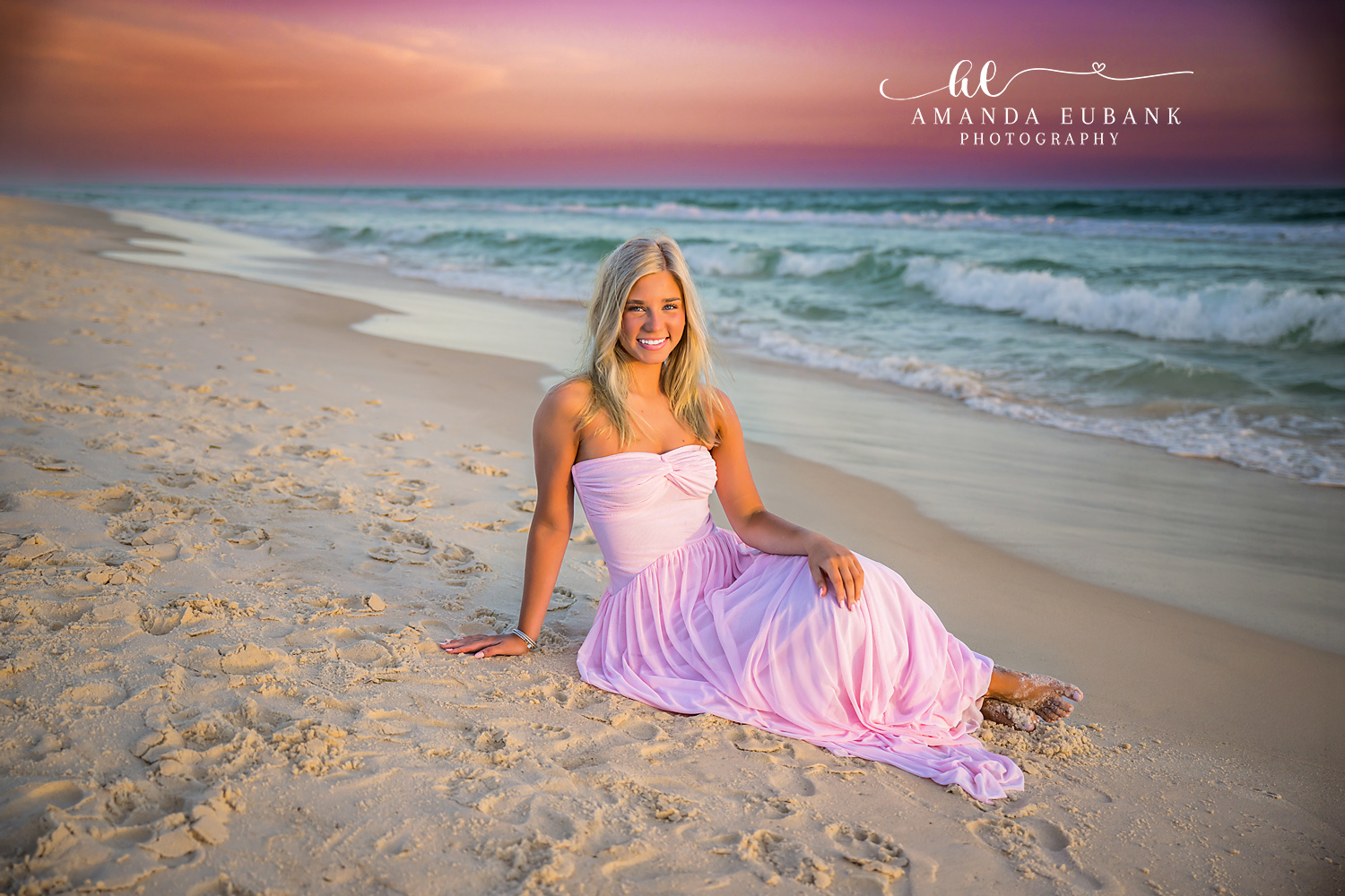 Rosemary Beach Pink Gown Beachfront Senior Portrait | 30A Senior Photographer Senior girl in a flowing blush pink gown sitting on the white sand at Rosemary Beach with turquoise Gulf water and a pastel sunset behind her