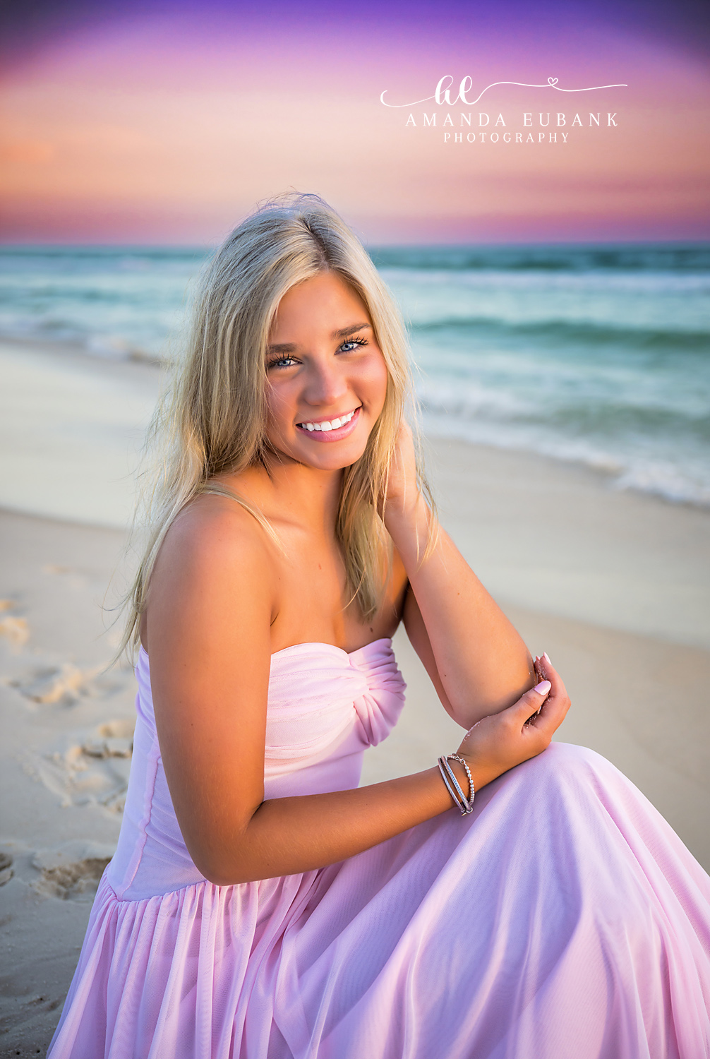 Rosemary Beach Pastel Sunset Senior Portrait | 30A Senior Photographer Senior girl in a blush pink gown kneeling on Rosemary Beach FL sand beneath a dreamy purple, pink, and teal pastel sunset sky