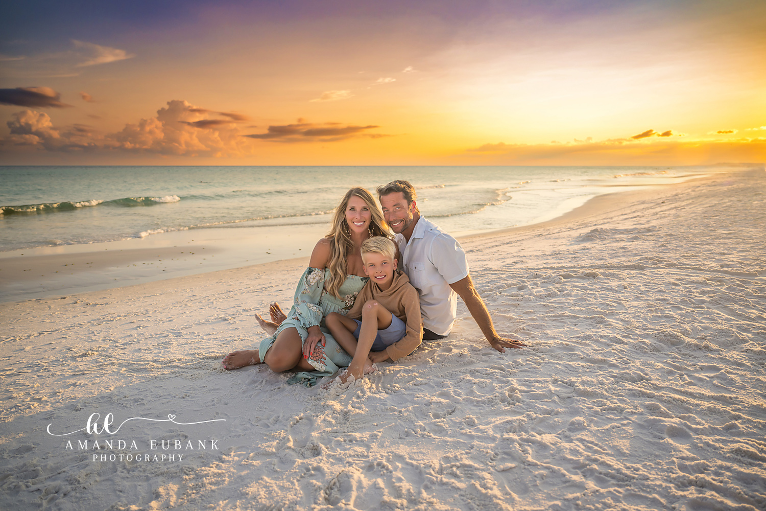Destin Beach Family Session – Family of Three at Golden Hour Family of three sitting together in the sand and smiling at sunset during a Destin beach family photography session