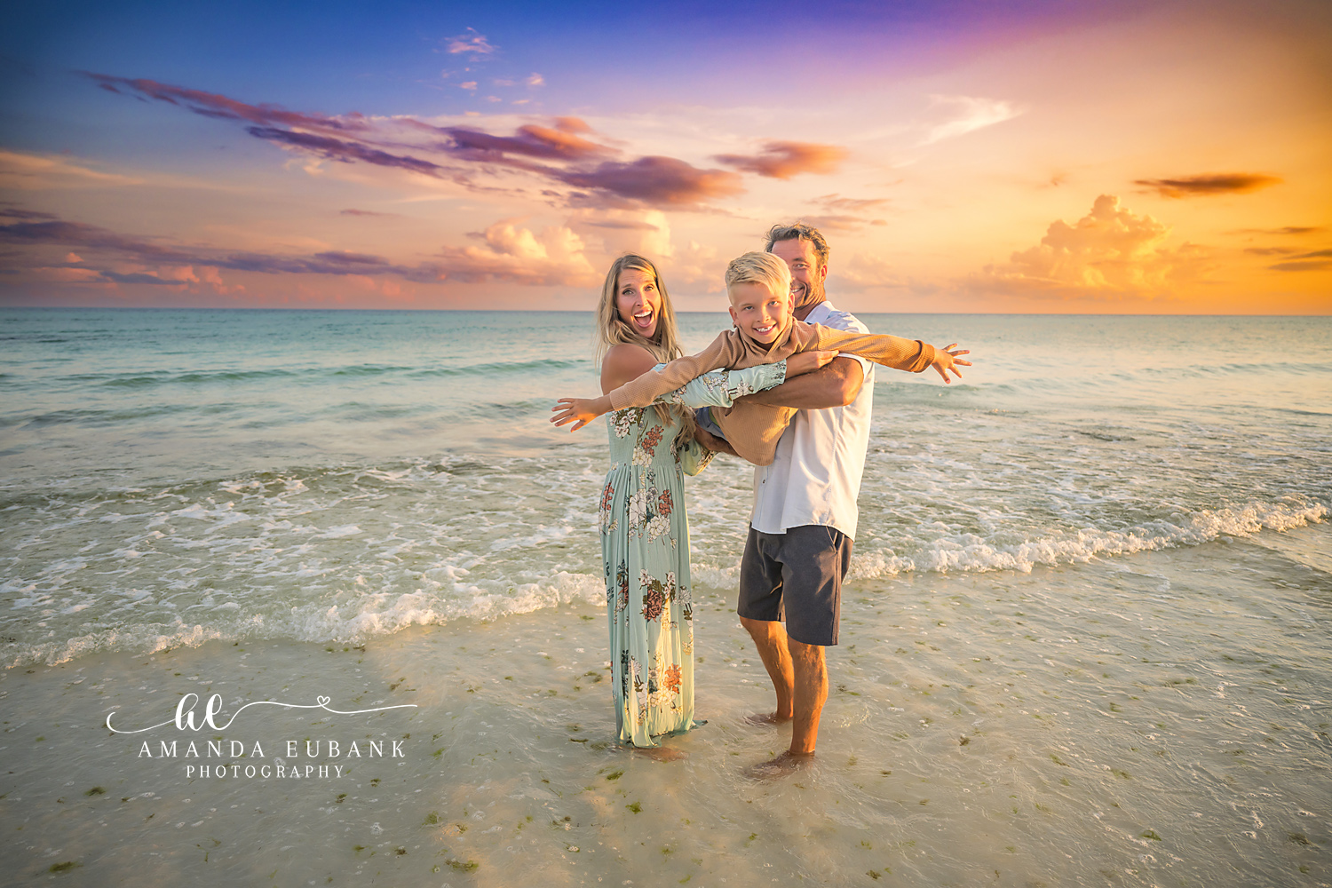 Father lifting his laughing son while mom smiles beside them on the shoreline at sunset in Destin