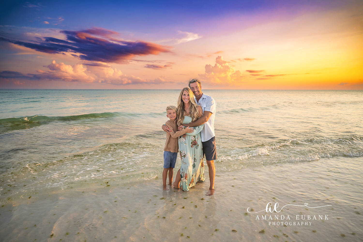 Destin Beach Family Session – Family Hug at Sunset Family of three hugging together at the water's edge under a golden sunset during a Destin beach family photography session