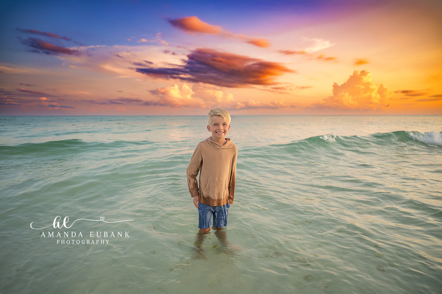 Destin Beach Family Session – Boy in the Gulf at Sunset Young boy standing waist deep in the Gulf beneath dramatic sunset clouds during a Destin beach family photography session