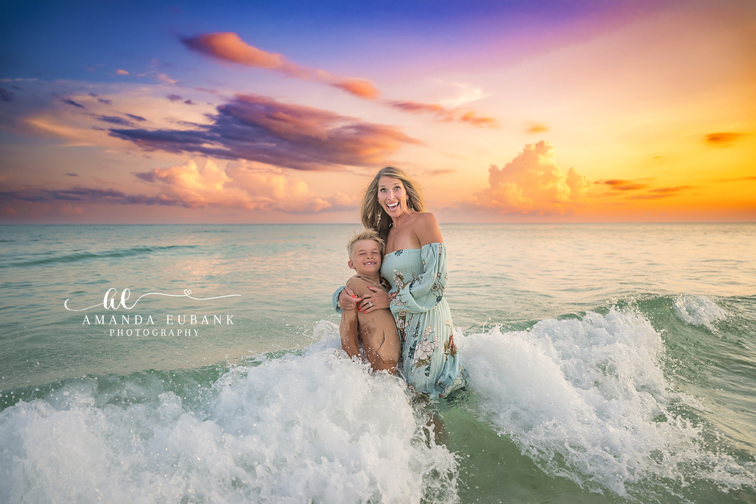 Destin Beach Family Session – Mother and Son in the Waves Mother in a floral dress holding her son as a wave breaks around them at sunset during a Destin family photography session