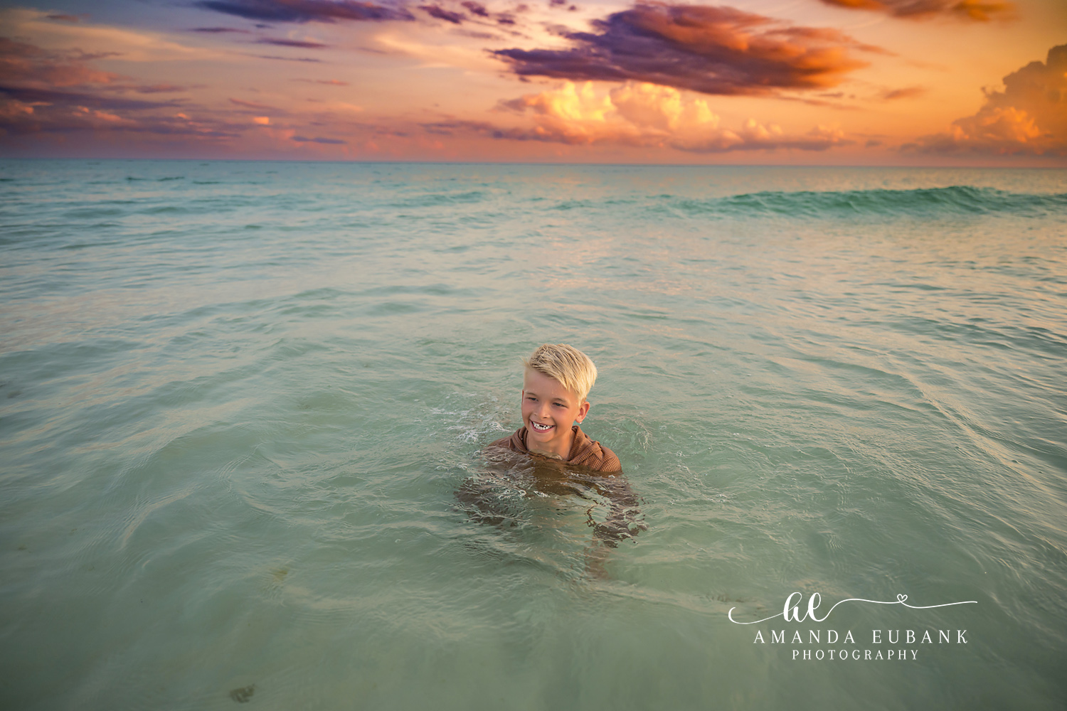 Destin Beach Family Session – Swimming in the Emerald Water Young boy swimming in the clear emerald Gulf water beneath a dramatic sunset sky during a Destin family photography session