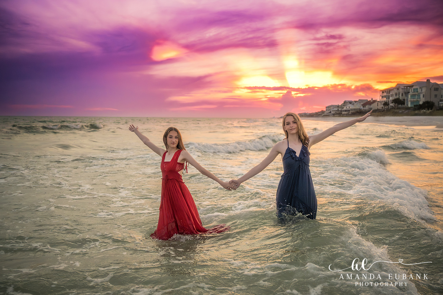 Rosemary Beach Senior Photographer – Hand in Hand at Sunset Two senior girls in red and navy dresses holding hands in the ocean at sunset in Rosemary Beach, Florida