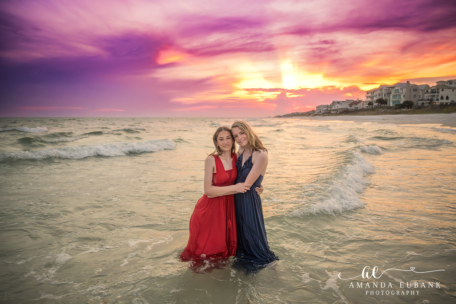 Rosemary Beach Senior Photographer – Sisters in the Surf at Sunset Two senior girls in red and navy dresses embracing in the ocean at sunset in Rosemary Beach, Florida
