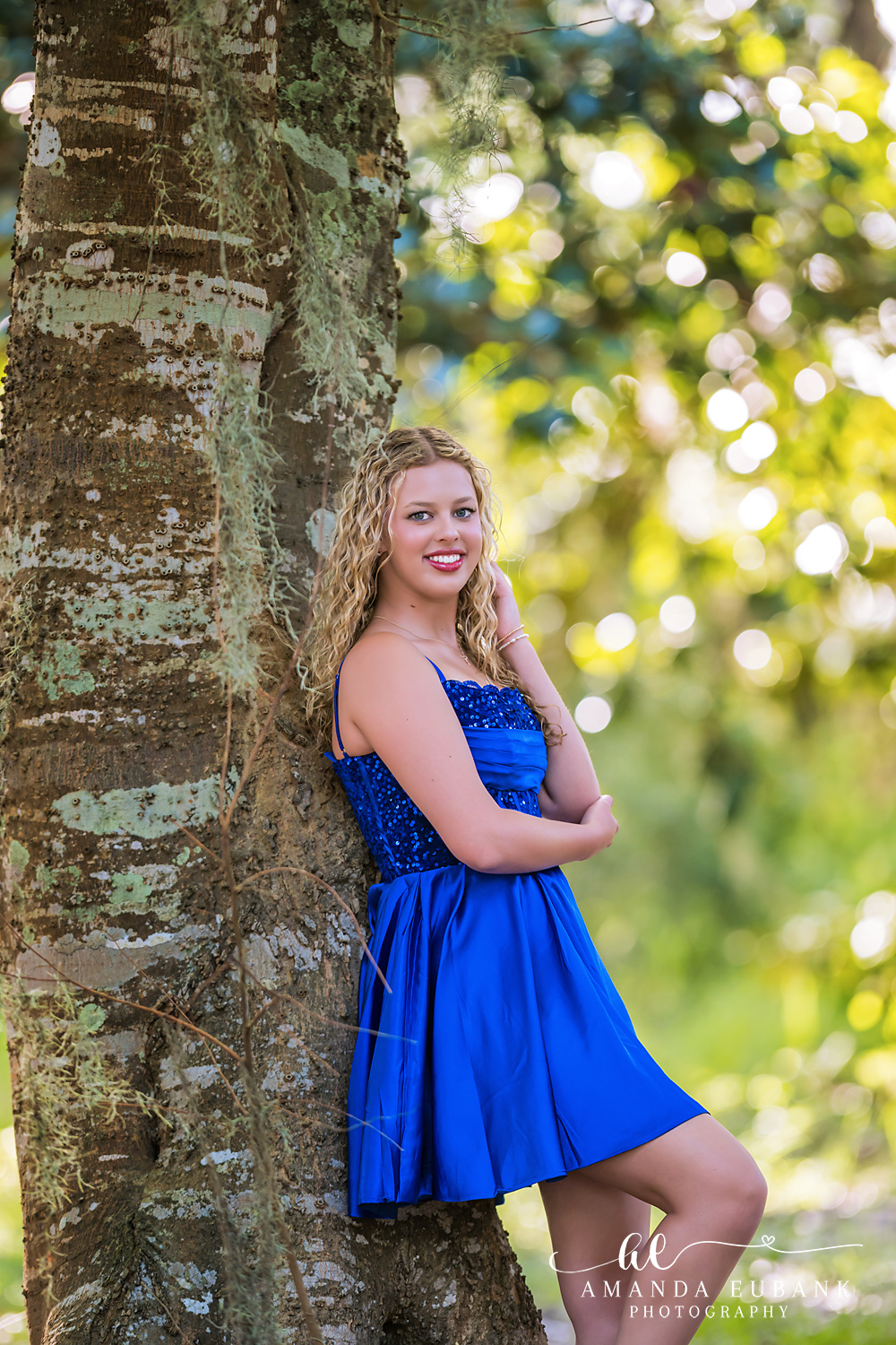 Sandestin Senior Photographer – Blue Dress Portrait Under the Oaks High school senior in a royal blue dress leaning against a tree during a Sandestin, Florida senior session