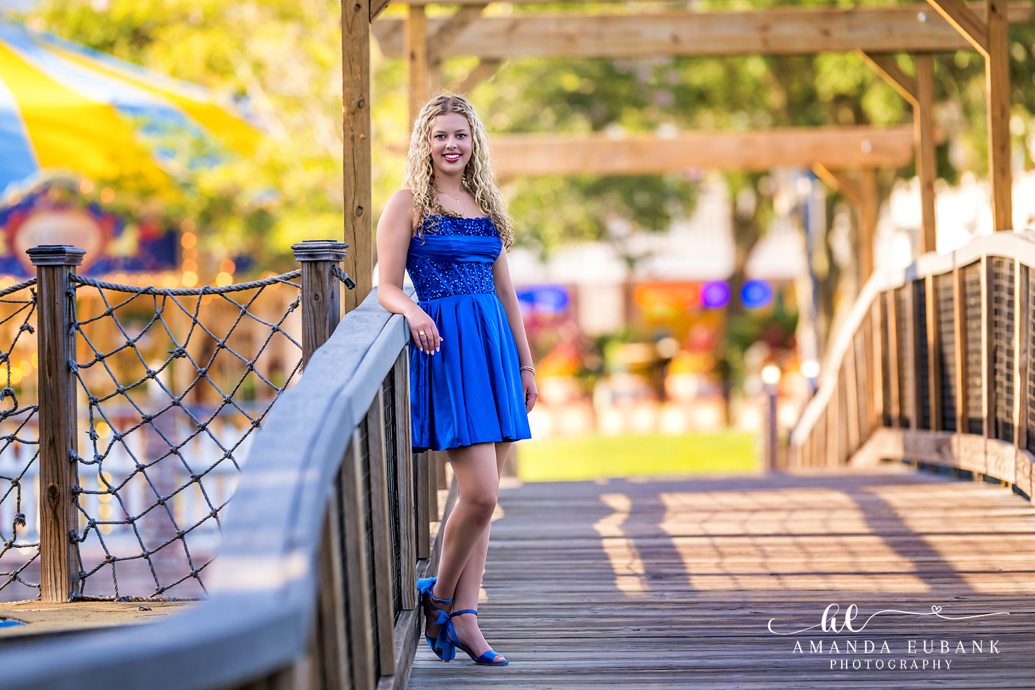 Sandestin Senior Photographer – Evening Portrait at Baytowne Wharf Senior in a blue dress posing on a bridge railing with string lights in the background in Sandestin, Florida