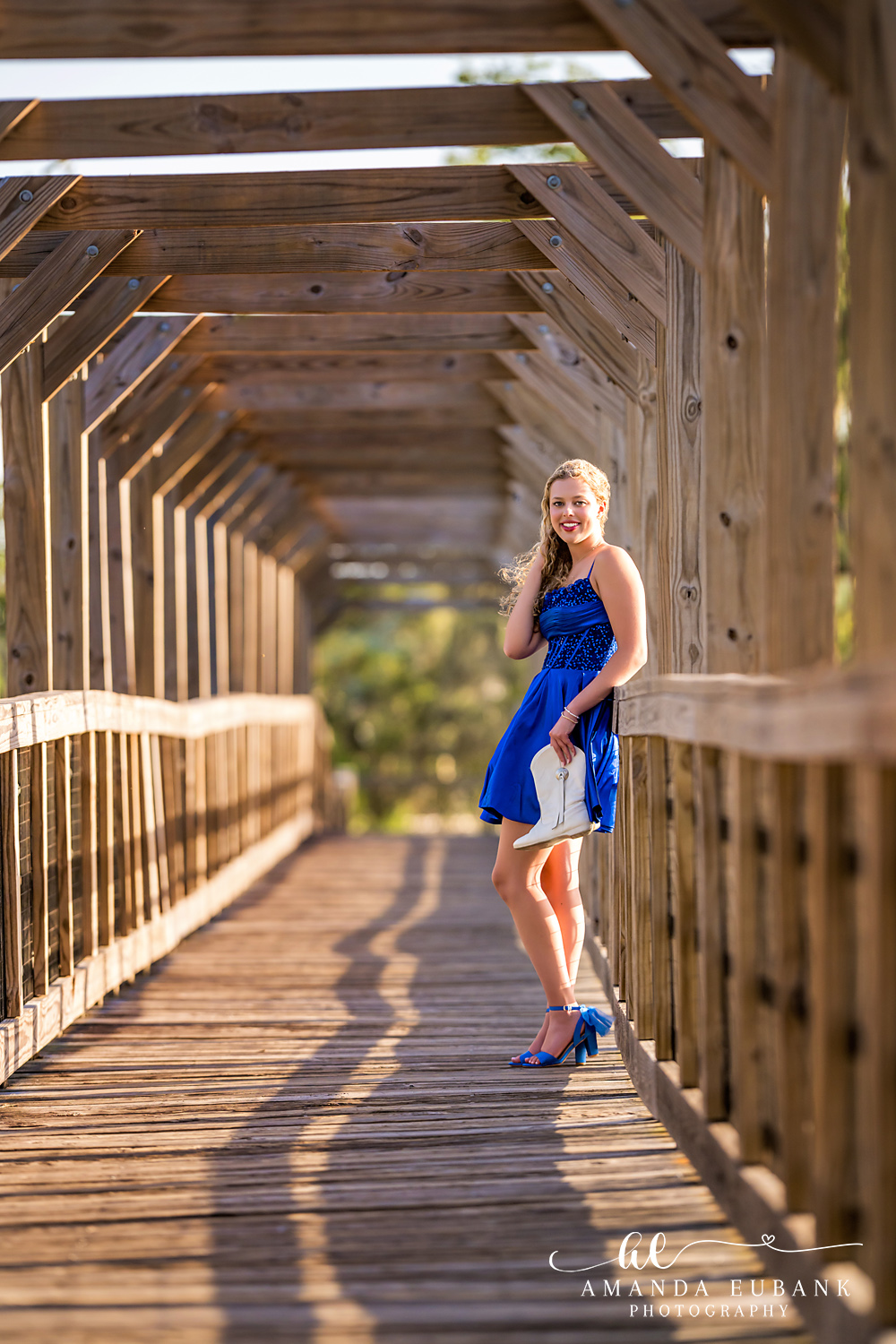 Sandestin Senior Photographer – Wooded Boardwalk Path Portrait High school senior walking a shaded wooden boardwalk path in a blue dress in Sandestin, Florida
