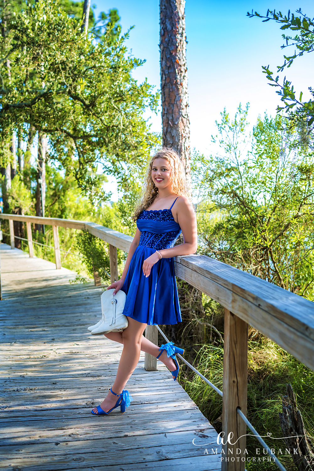 Sandestin Senior Photographer – Bridge Railing Portrait in the Woods Senior in a blue dress leaning on a wooden bridge railing surrounded by trees in Sandestin, Florida