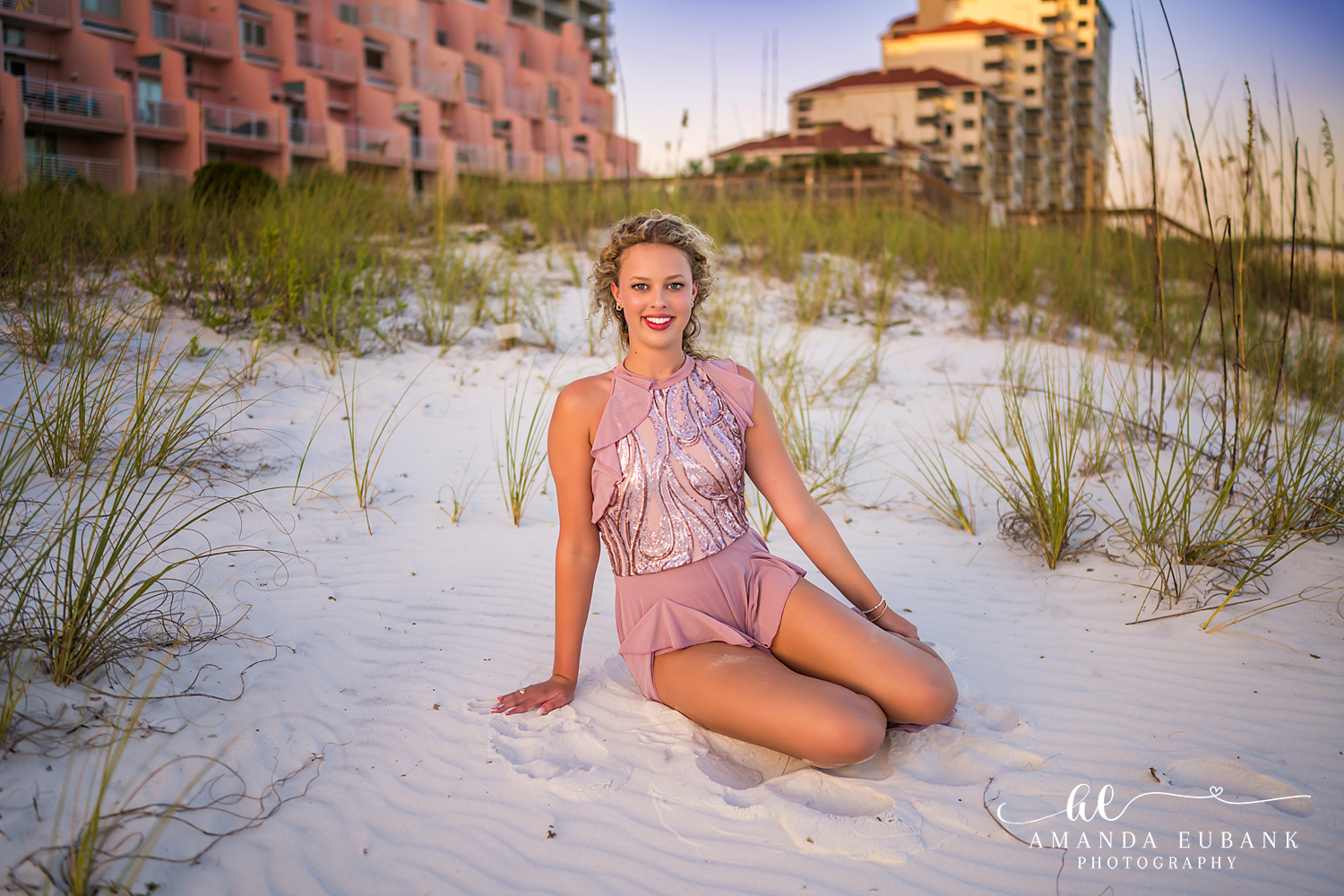 Sandestin Senior Photographer – Smiling Portrait in the Dunes High school senior in a pink dress smiling among the sand dunes with condos in the background in Sandestin, Florida