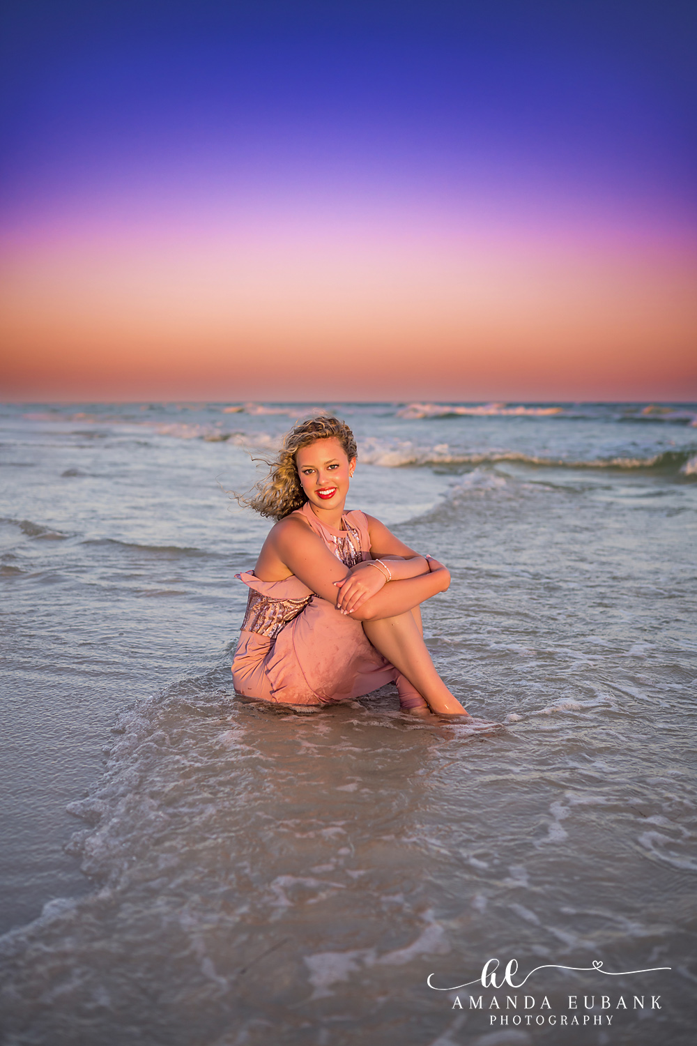Sandestin Senior Photographer – Seated at the Water’s Edge Senior girl sitting on the sand at the water's edge in a pink dress during sunset in Sandestin, Florida