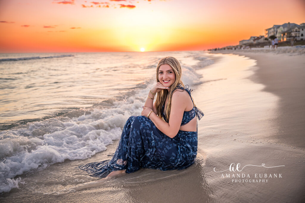 High school senior in a navy floral gown sitting in the shallow surf at sunset during a Santa Rosa Beach senior photography session