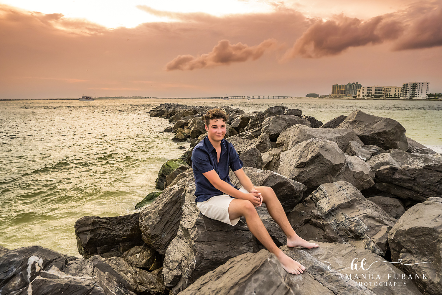 Destin Jetties Family Photos | Teen boy sitting A teen boy sitting on the rocks at the Destin Jetties in Destin, Florida at sunset, photographed by Destin family photographer Amanda Eubank