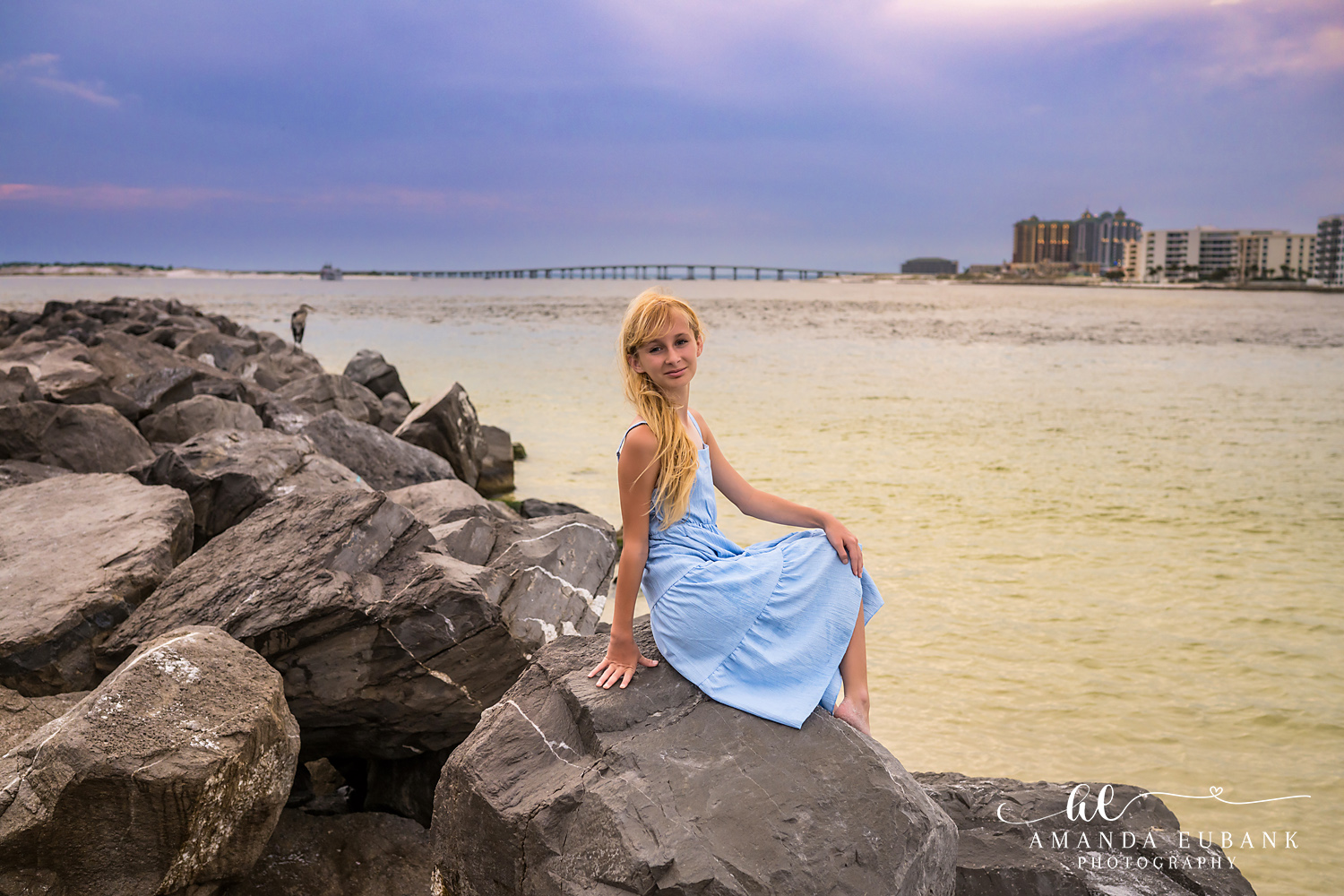 Destin Jetties Family Photos | Girl in a blue dress seated A girl in a blue dress seated on the rocks at the Destin Jetties in Destin, Florida at sunset, photographed by Destin family photographer Amanda Eubank
