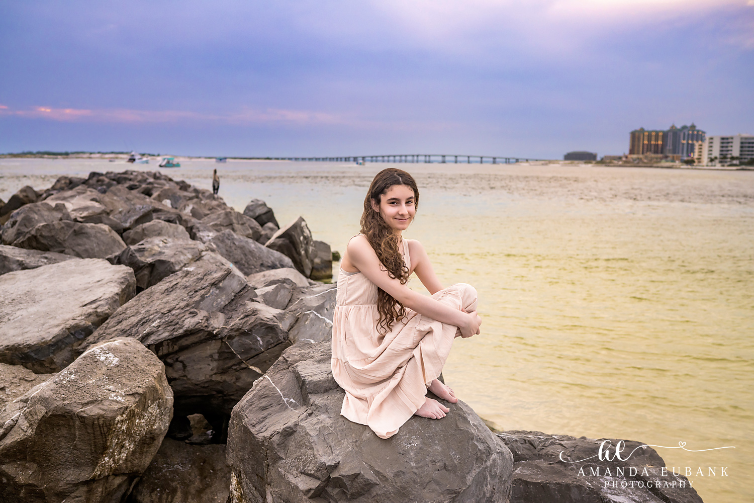 Destin Jetties Family Photos | Girl in a blush dress seated A girl in a blush dress seated on the rocks at the Destin Jetties in Destin, Florida at sunset, photographed by Destin family photographer Amanda Eubank