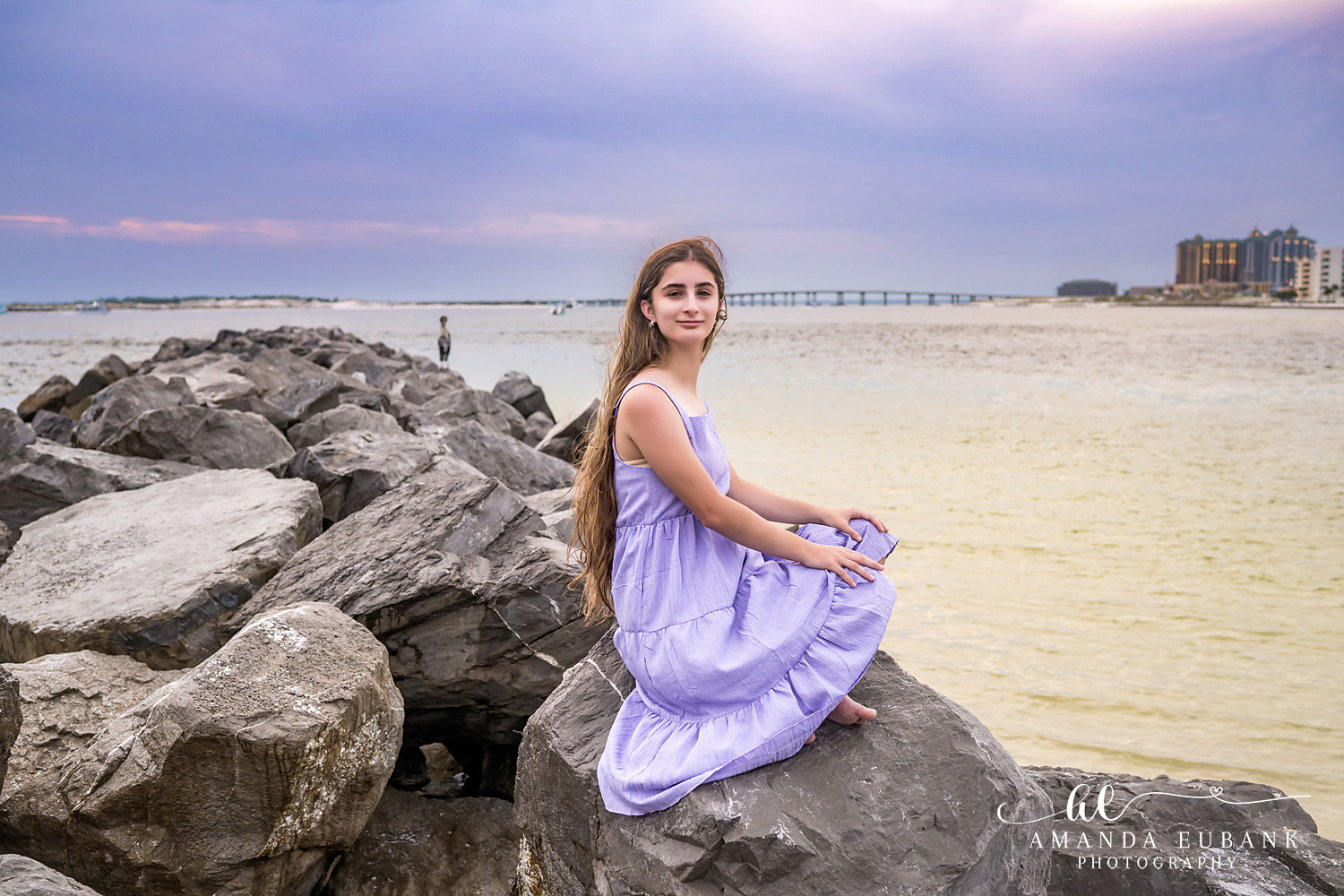Destin Jetties Family Photos | Girl in a lavender dress seated A girl in a lavender dress seated on the rocks at the Destin Jetties in Destin, Florida at sunset, photographed by Destin family photographer Amanda Eubank