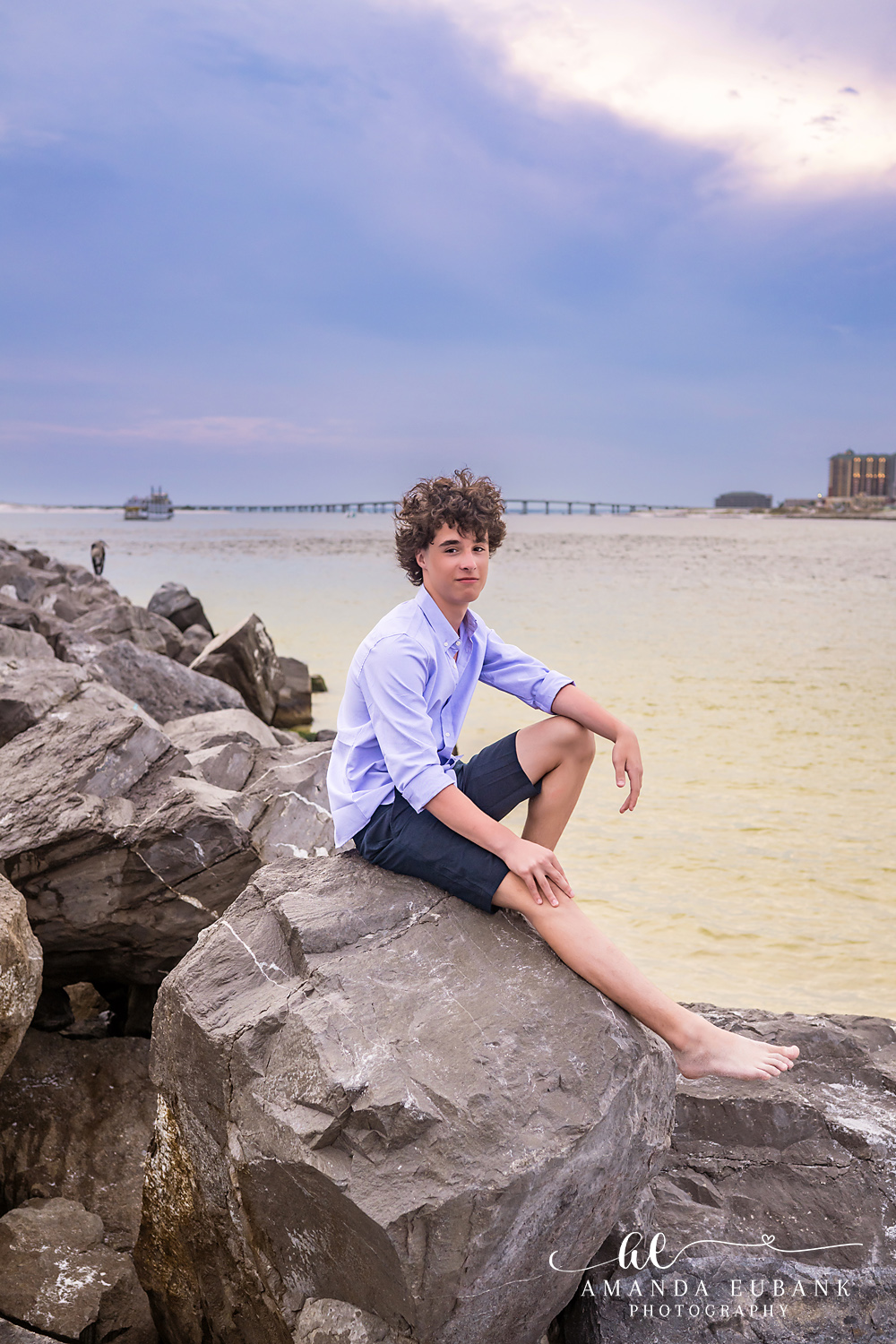 Destin Jetties Family Photos | Teen boy sitting barefoot A teen boy sitting barefoot on the rocks at the Destin Jetties in Destin, Florida at sunset, photographed by Destin family photographer Amanda Eubank