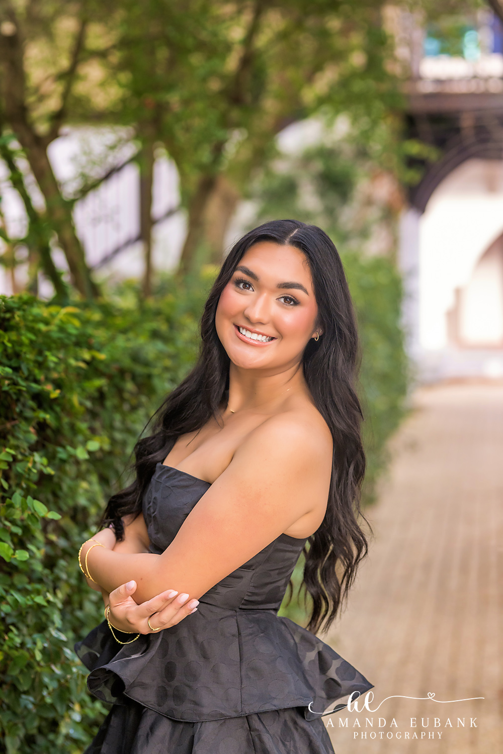 Rosemary Beach Senior Photos | Black tiered dress on a brick walkway Rosemary Beach senior photographer: senior girl in a black strapless tiered dress smiling with arms folded on a brick walkway lined with greenery