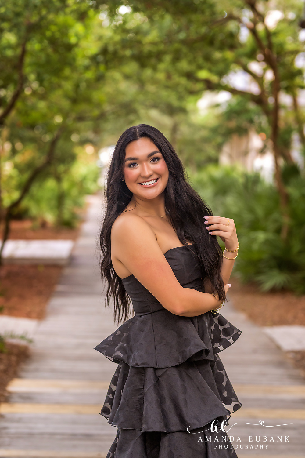 Rosemary Beach Senior Photos | Standing on a boardwalk path Rosemary Beach senior photographer: senior girl in a black tiered dress smiling with hand on her shoulder on a wooden boardwalk lined with trees