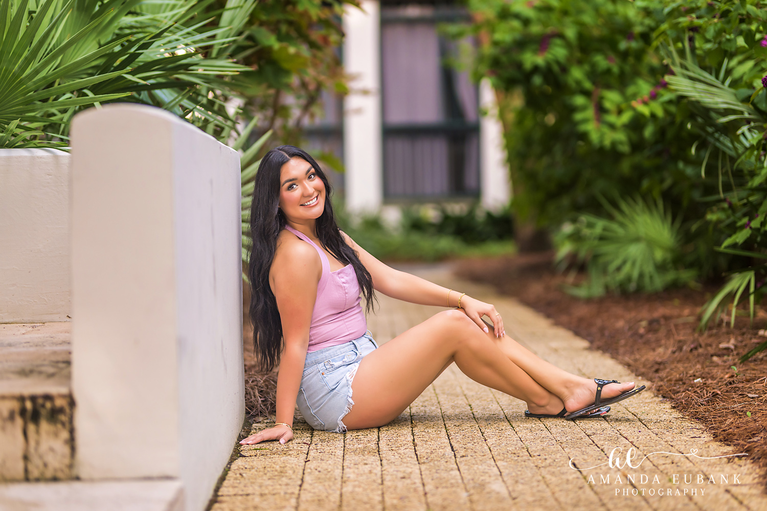 Rosemary Beach Senior Photos | Seated on a brick path Rosemary Beach senior photographer: senior girl in a pink top and denim shorts seated on a brick path against a white wall with tropical plants