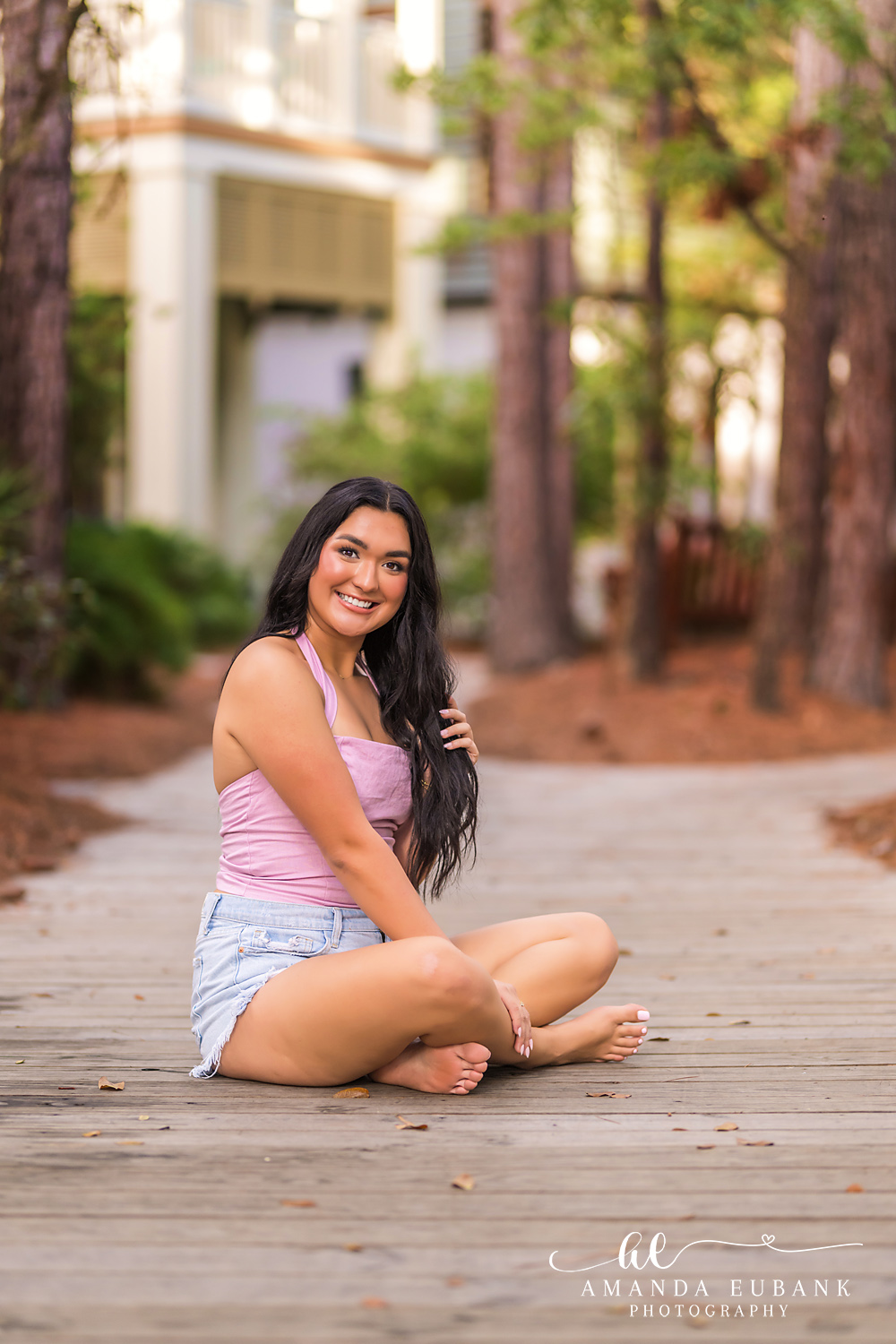 Rosemary Beach Senior Photos | Seated on a wooden boardwalk Rosemary Beach senior photographer: senior girl in a pink halter top and denim shorts sitting cross legged on a wooden boardwalk among pine trees