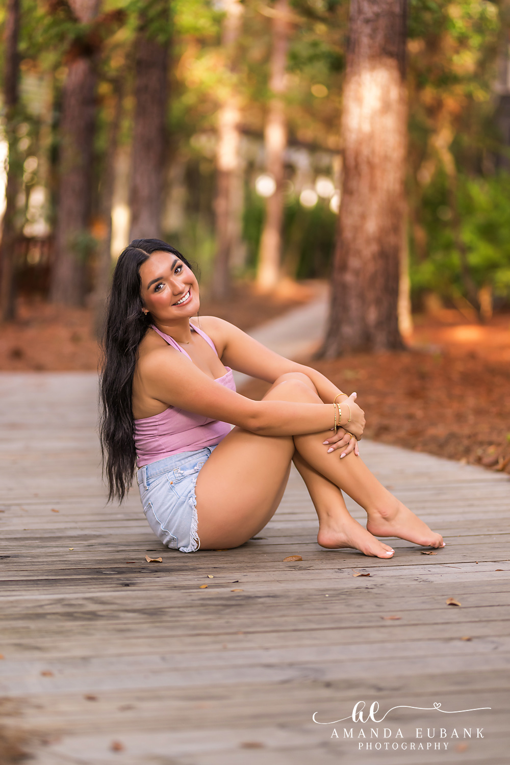 Rosemary Beach Senior Photos | Hugging her knees on the boardwalk Rosemary Beach senior photographer: senior girl in a pink top and denim shorts sitting and hugging her knees on a wooden boardwalk in a pine grove